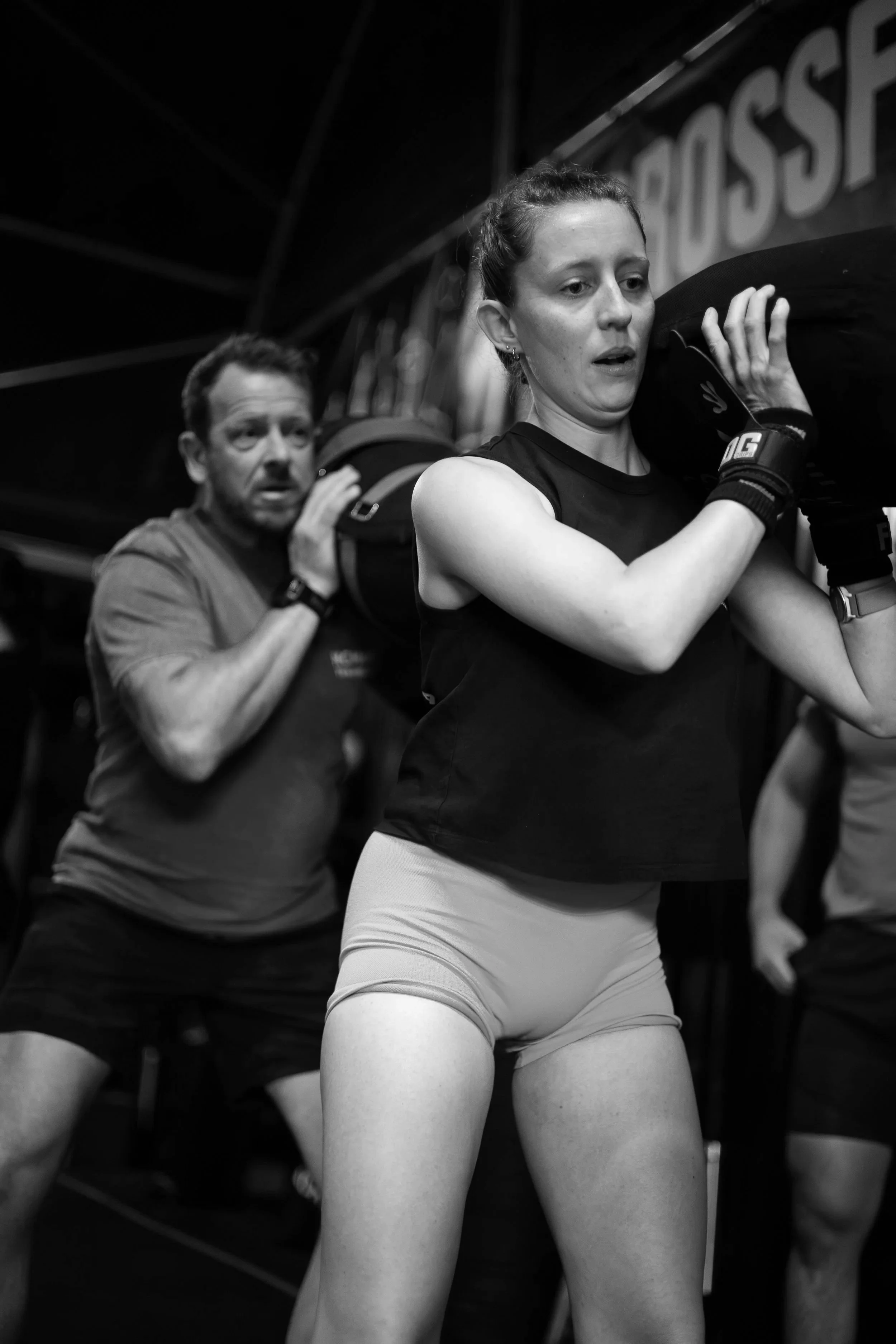 A woman lifting a weight with her shoulders during a workout, with a man in the background also lifting a weight.
