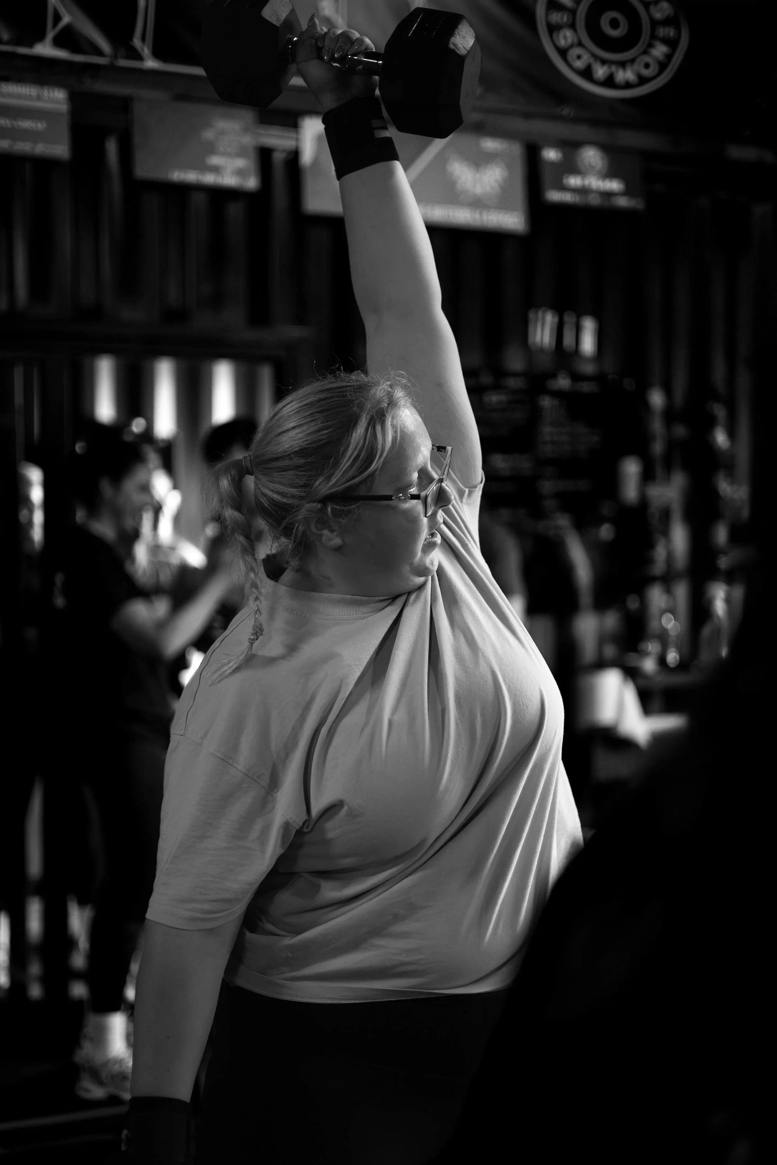 A woman lifting a dumbbell in a gym, wearing glasses and a light-colored t-shirt.