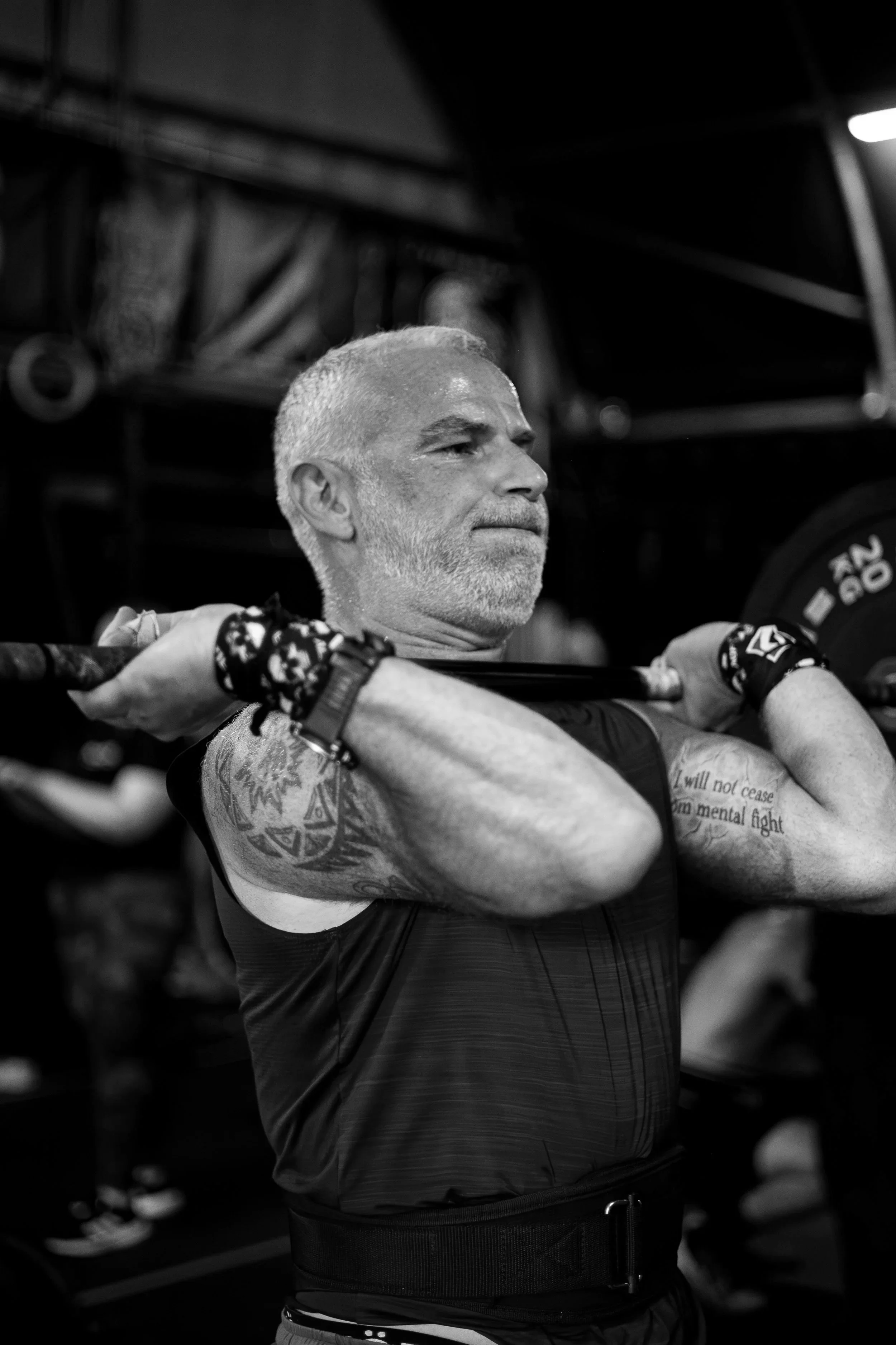 A middle-aged man with gray hair and beard lifting a barbell in a gym, with muscular arms and tattoos visible.