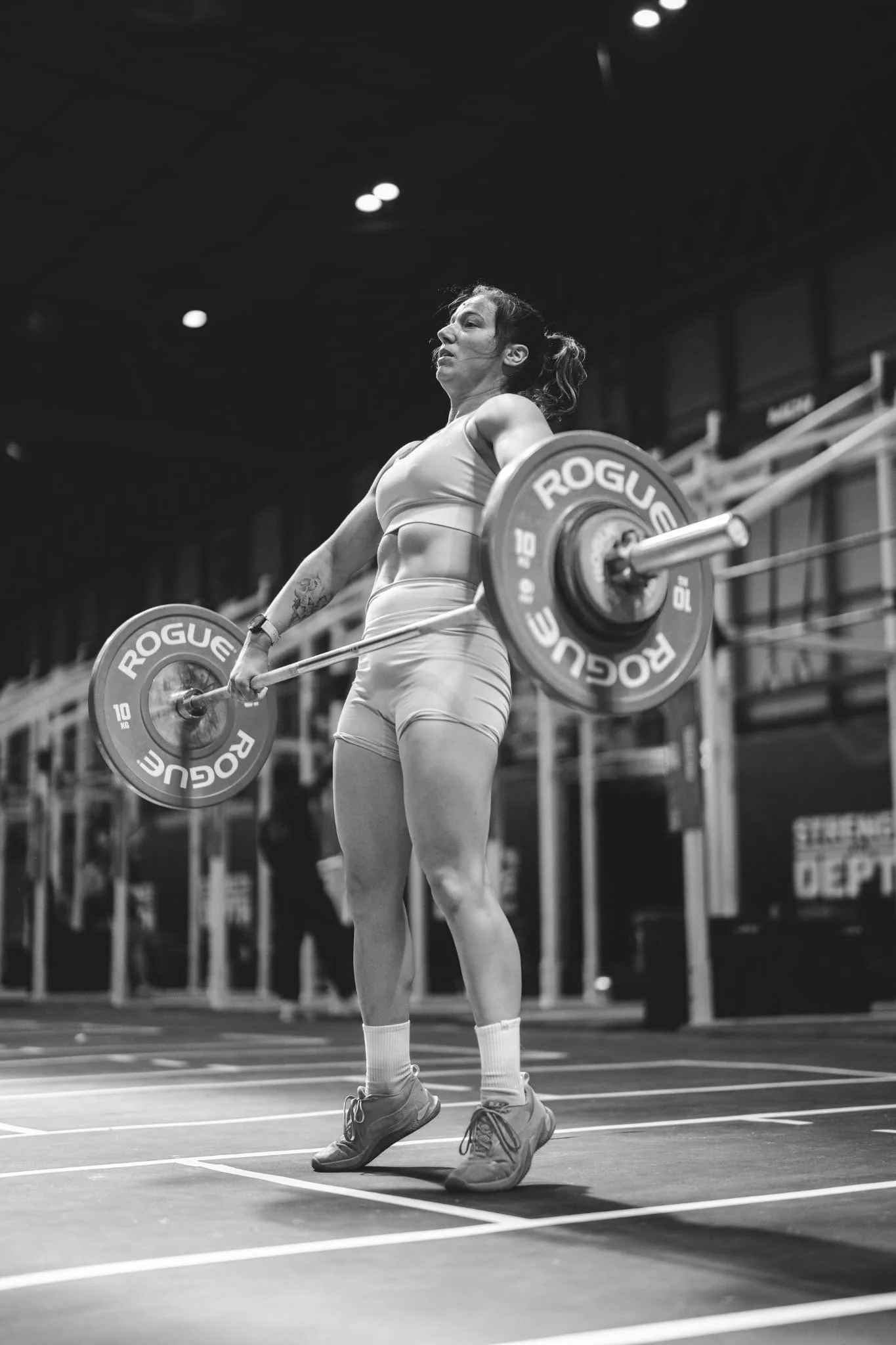A woman with a ponytail lifting a barbell at an indoor sports facility. She wears athletic clothes and sneakers, and stands on a court with marked lines. The background shows seating and some equipment, and there's a dark ceiling with lights.