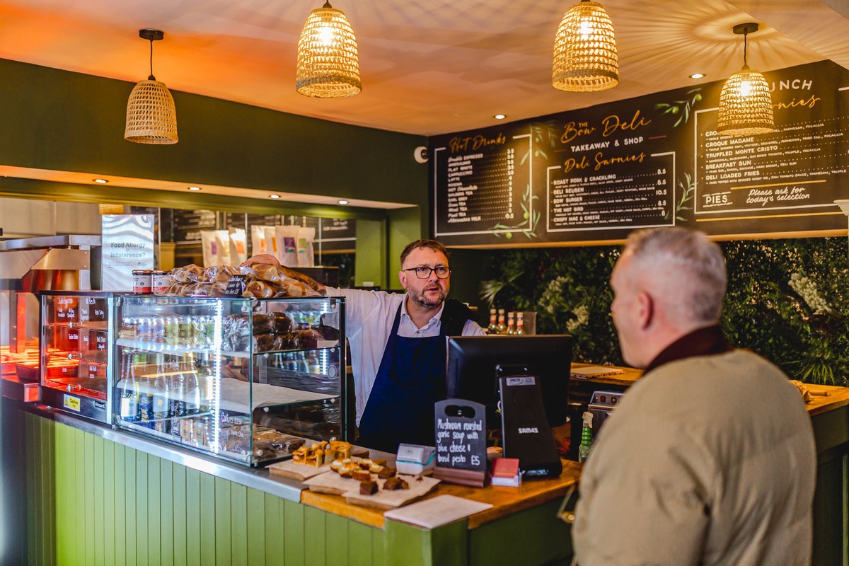 Man behind a counter in a deli talking to a customer, with food and drinks displayed, and menu boards on the wall.