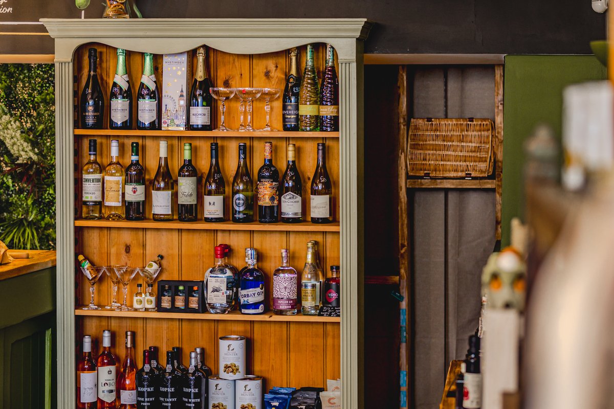 A wooden shelf stocked with various bottles of wine, spirits, and champagne. Some cocktail glasses and boxes are also displayed. The shelf is in a rustic setting with plants nearby.