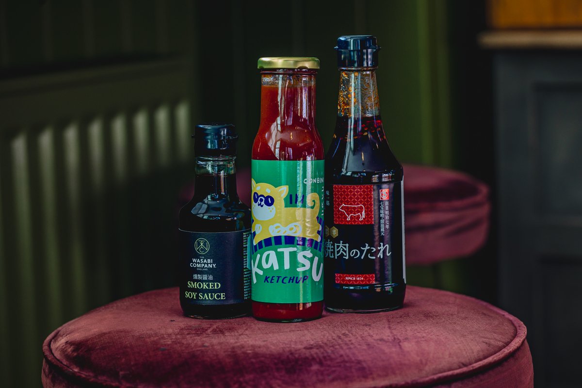 Three condiment bottles on a velvet stool; smoked soy sauce on the left, a bottle labeled 'Katsu Ketchup' in the center, and a bottle with Asian characters on the right.