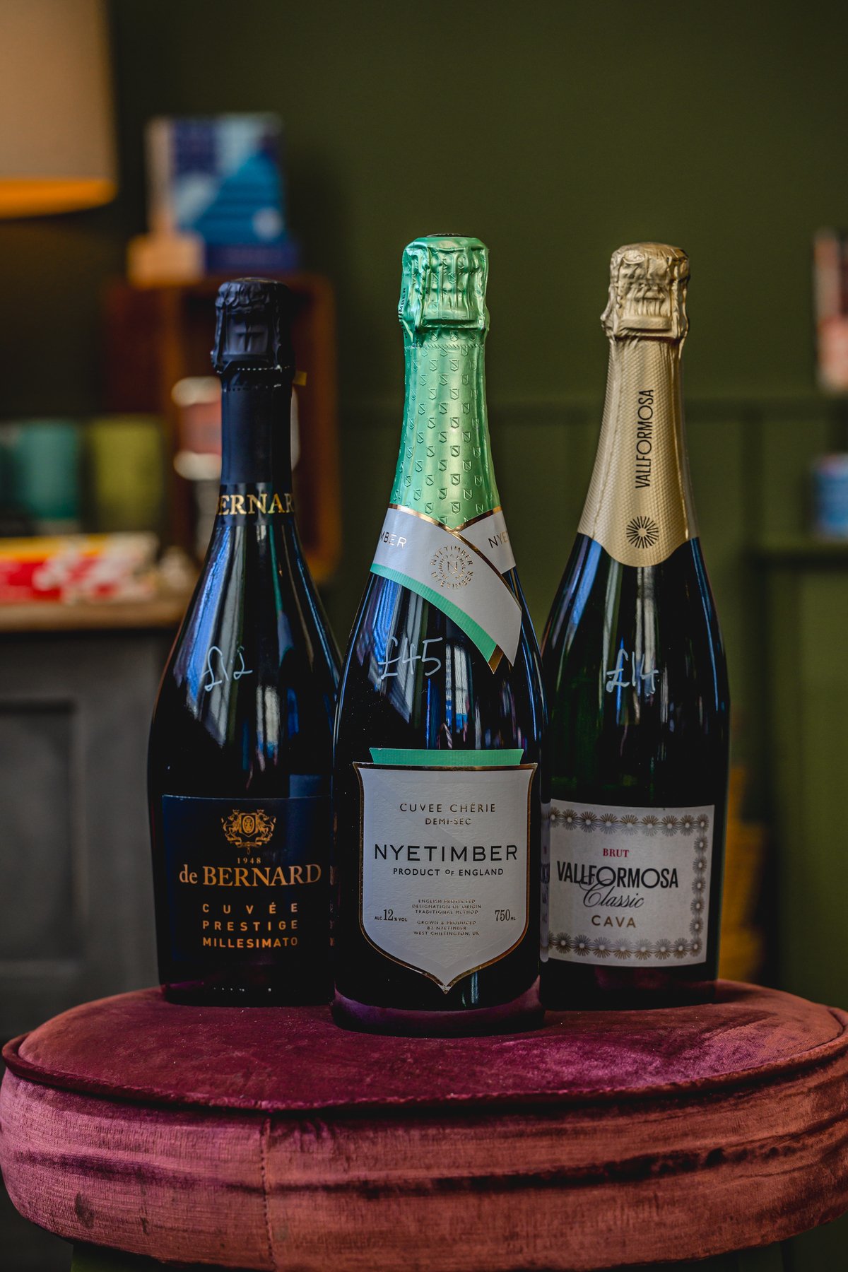 Three bottles of sparkling wine on a red velvet cushion, featuring de Bernard, Nyetimber, and Vallformosa brands, with green and gold foil tops.