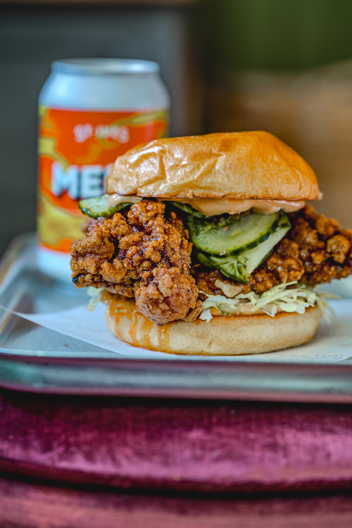 Spicy fried chicken sandwich with pickles on a tray next to a colorful beverage can.