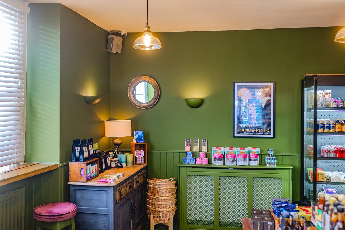 Cozy interior of a store with green walls, featuring a wooden sideboard displaying various packaged products, a lamp, decorative mirror, and a refrigerator stocked with beverages. A poster hangs on the wall and woven baskets are stacked on the floor.