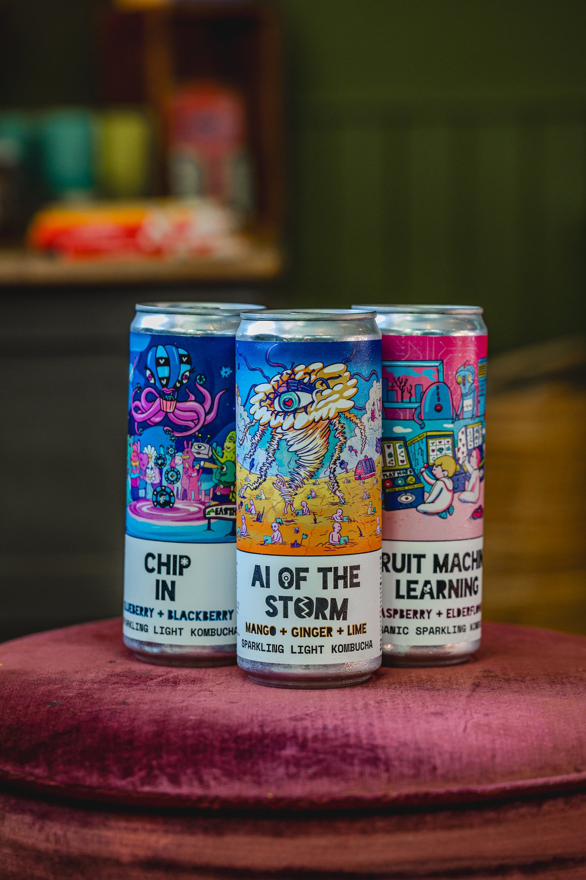 Three cans of kombucha with colorful, artistic labels displayed on a fabric surface. The flavors are "Chip In" with blueberry and blackberry, "AI of the Storm" with mango, ginger, and lime, and "Fruit Machine Learning" with raspberry and elderflower.