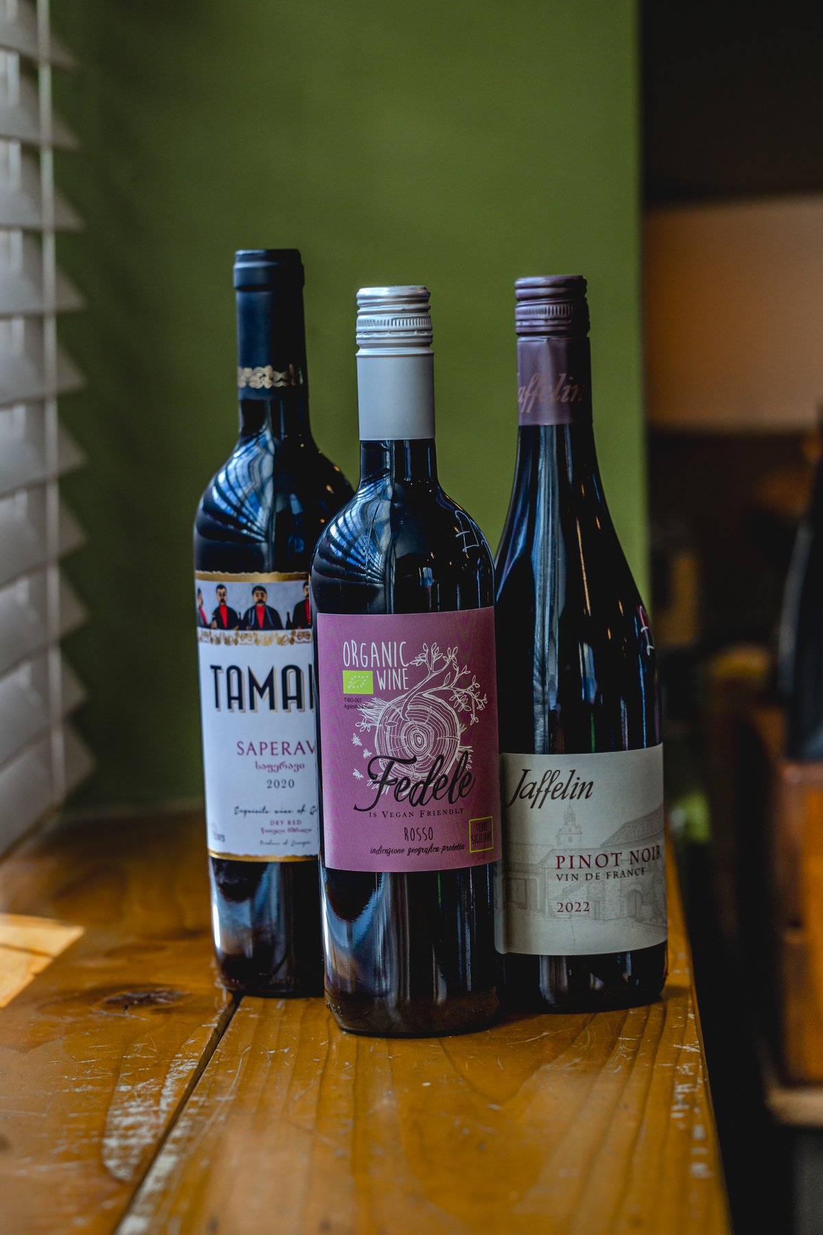 Three bottles of wine on a wooden table, featuring Tamada Saperavi 2020, Fedele Organic Vegan Friendly Rosso, and Jaffelin Pinot Noir 2022.