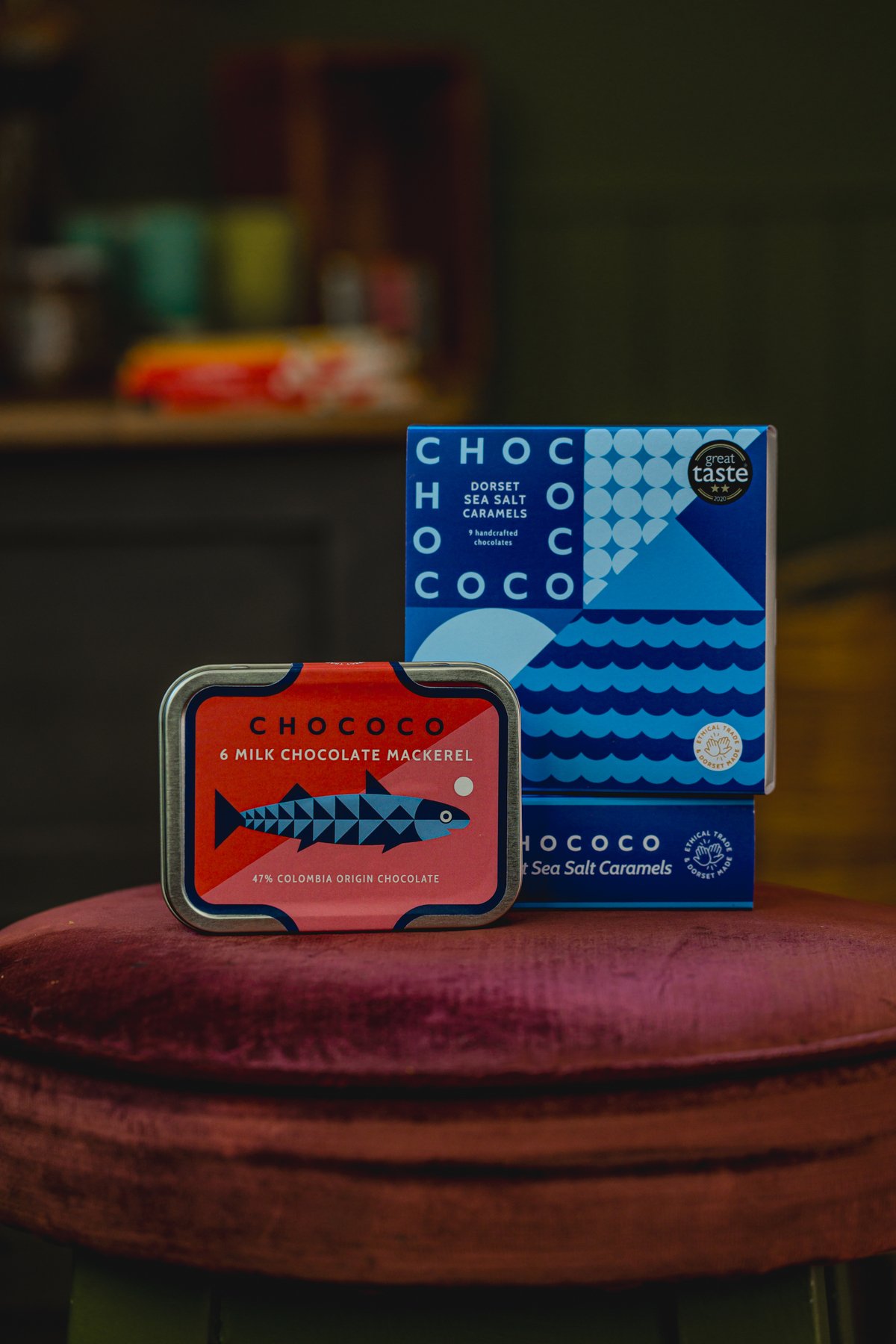Chococo chocolate products, including a tin of milk chocolate mackerel and a box of Dorset sea salt caramels, displayed on a maroon surface.