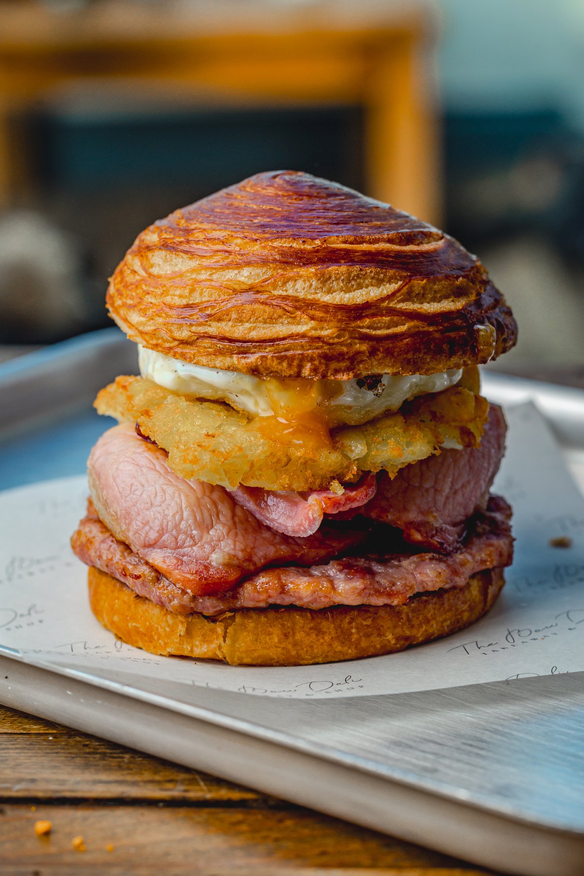 Gourmet breakfast sandwich with bacon, egg, and hash brown on a flaky croissant bun, served on a tray.