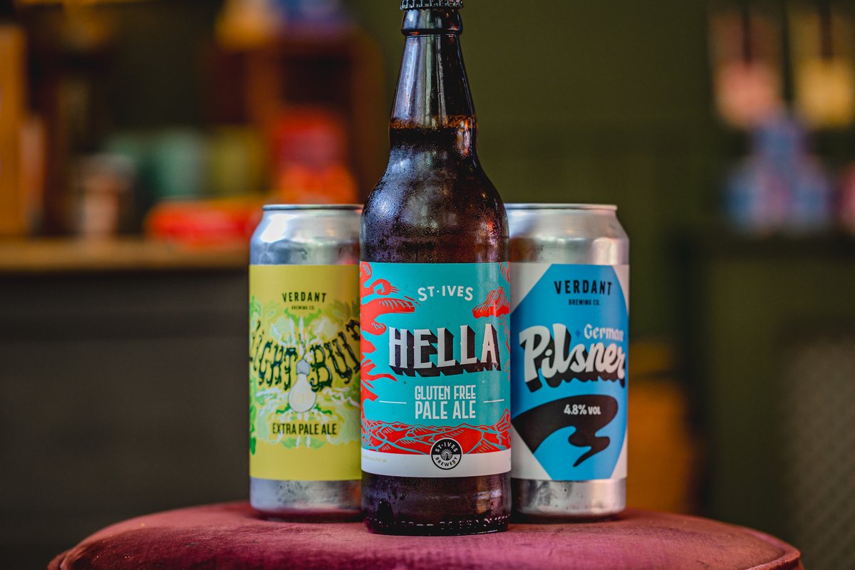 Three craft beer containers on a table; a bottle labeled "Hella Gluten Free Pale Ale St Ives" with vibrant red and teal colors, and two cans from Verdant Brewing Co - "Light Bulb Extra Pale Ale" with a yellow label, and "German Pilsner" with a blue l