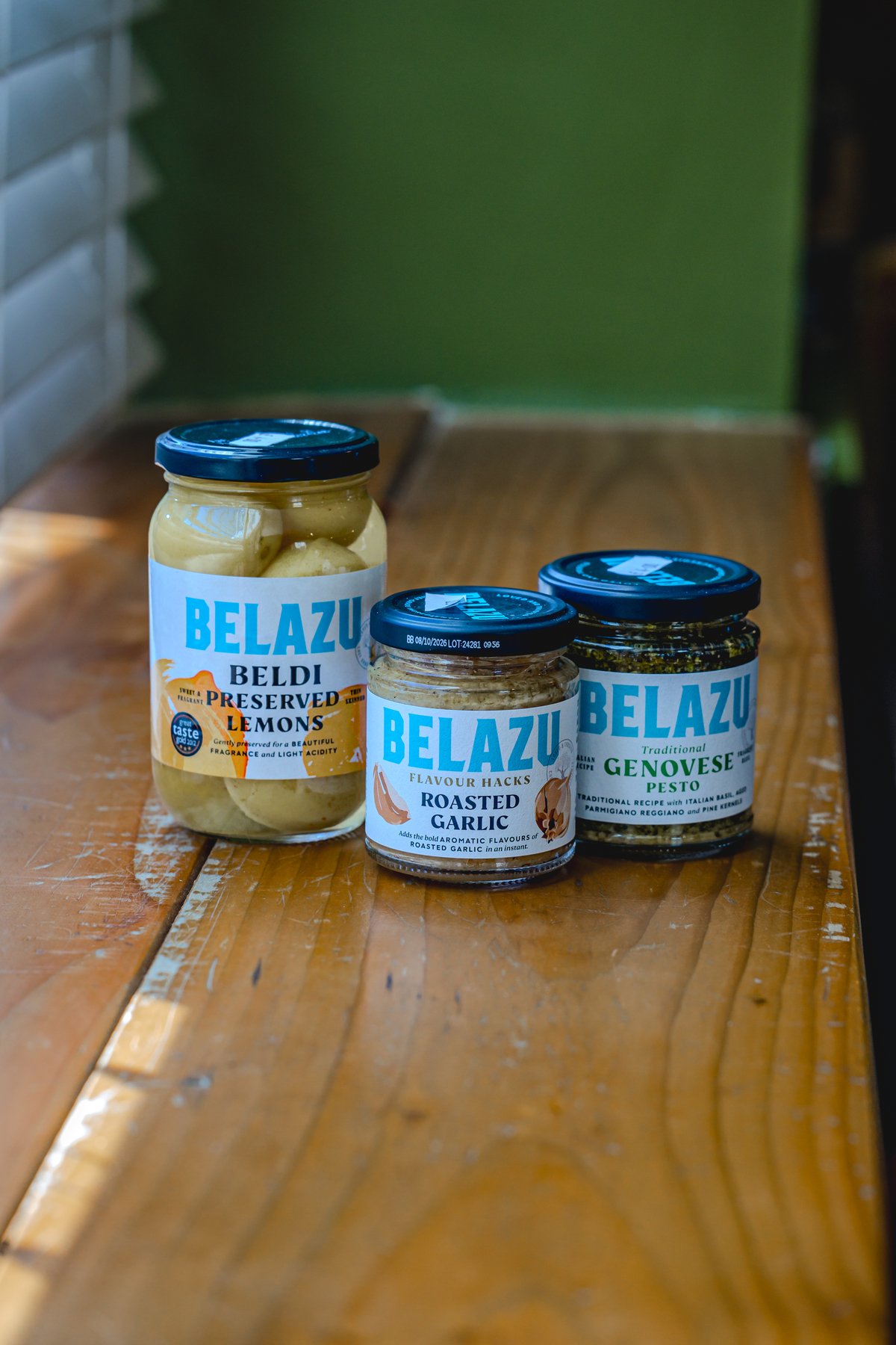 Three jars of Belazu products on a wooden table, including preserved lemons, roasted garlic, and Genovese pesto.