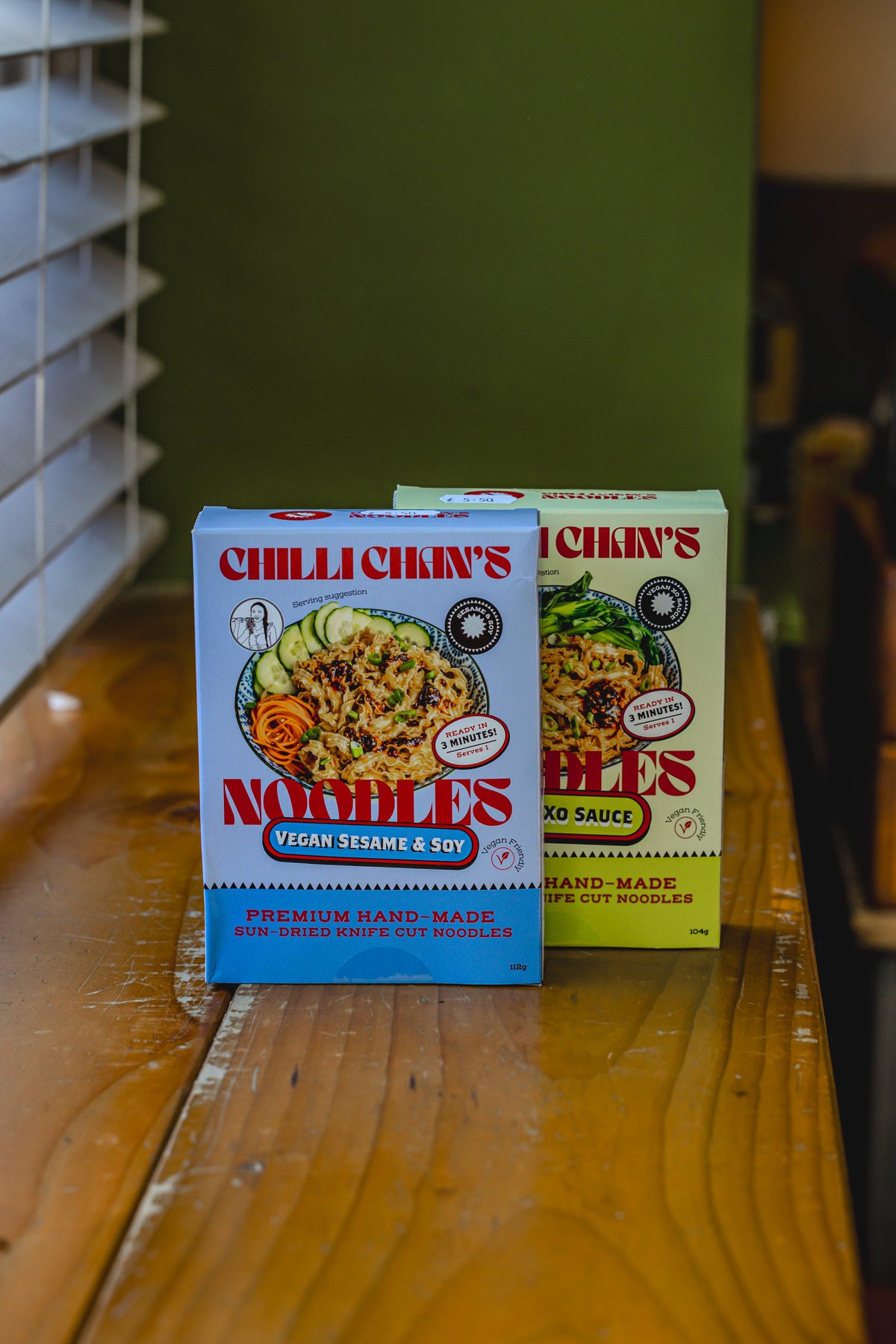 Two boxes of Chilli Chan's noodles on a wooden surface, one with vegan sesame & soy flavor, the other with XO sauce flavor.