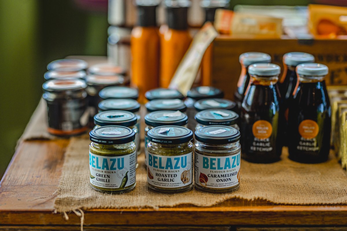Jars of Belazu brand condiments, including green chili, roasted garlic, and caramelized onion, arranged on a wooden table.