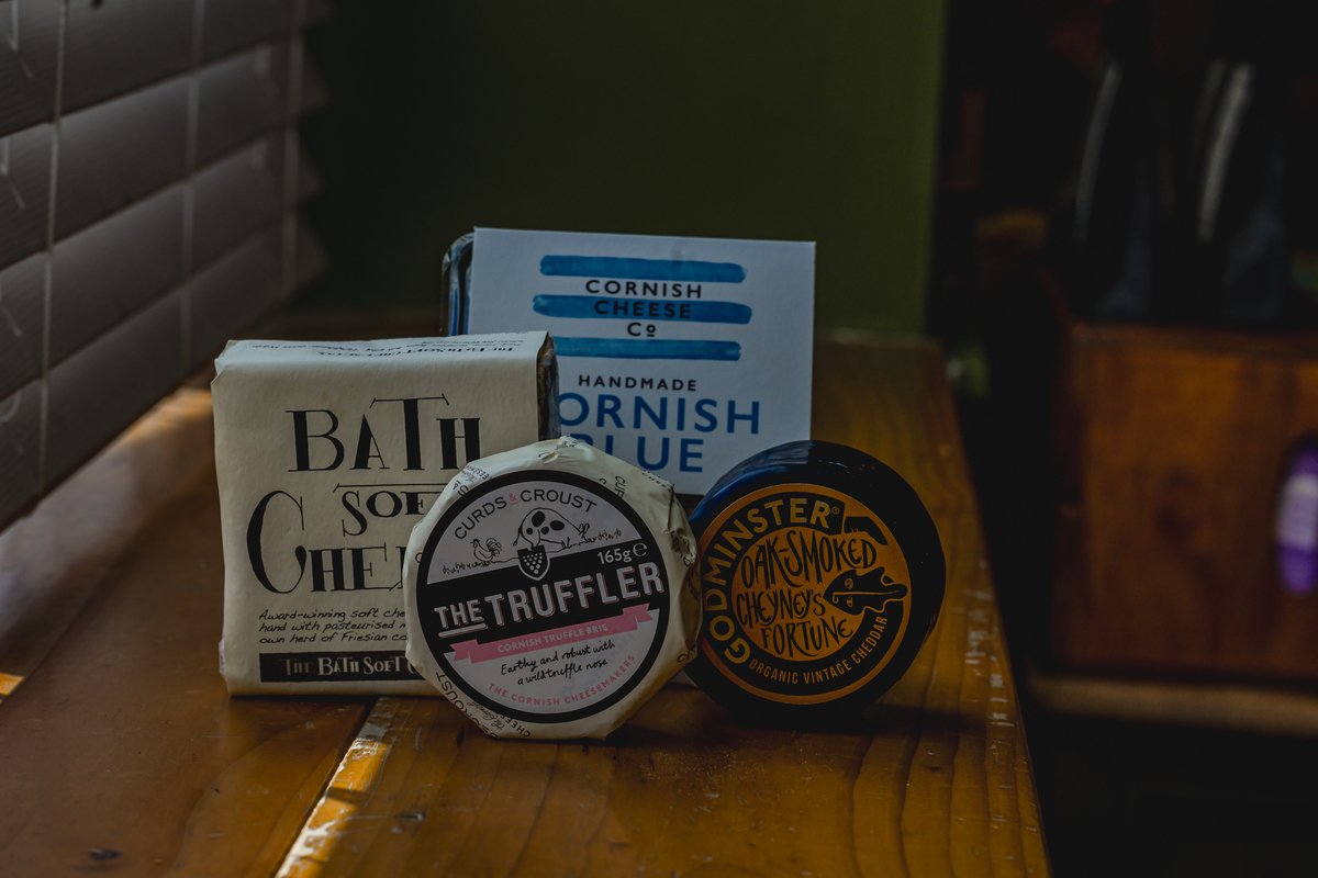 Variety of packaged cheeses on a wooden table, including Bath Soft Cheese, Cornish Blue Cheese, Truffled Brie, and Godminster Oak-Smoked Cheddar.