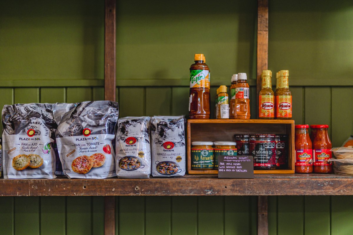 Assorted packaged food items on a wooden shelf, including bags of bread and paella rice from Plaza del Sol, Tajín seasoning, hot sauce bottles, jars of mole and chili paste, and salsa bottles.