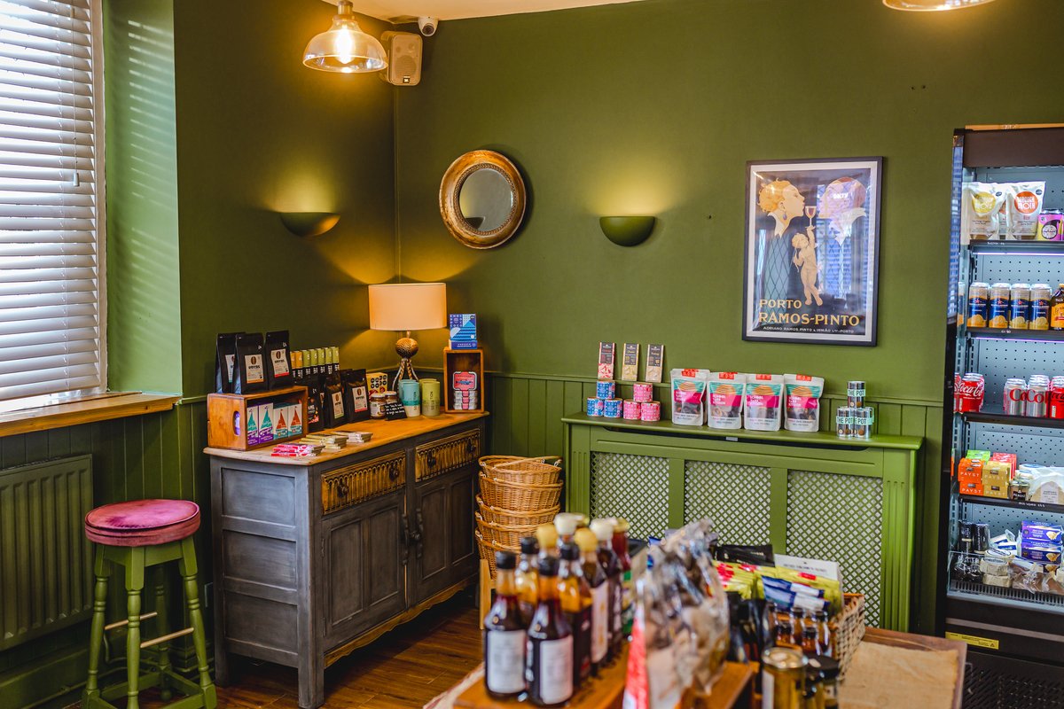 Cozy coffee shop interior with green walls, wooden furniture, snacks on display, a small refrigerator stocked with beverages, and decorative wall art.