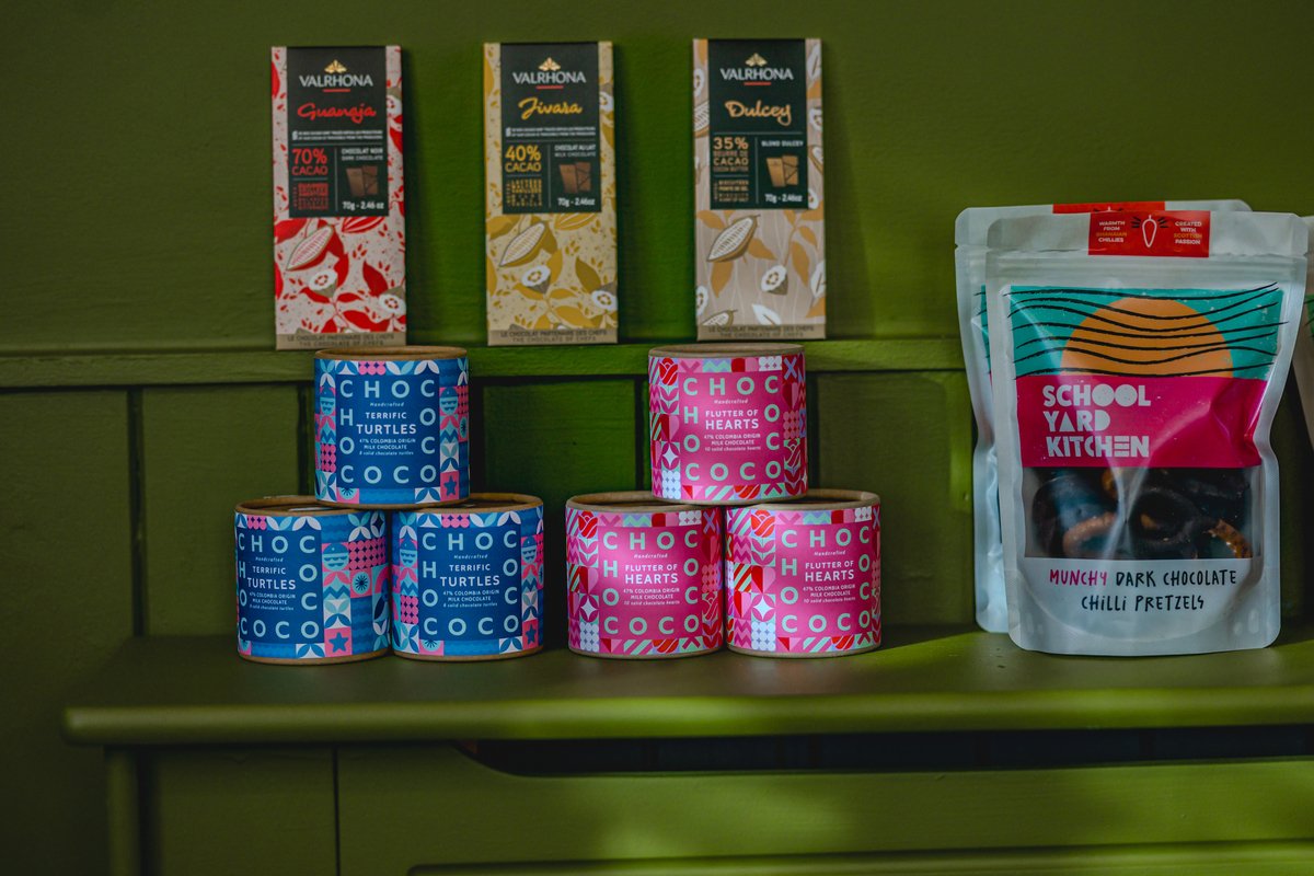Assorted chocolate products displayed against a green background. Includes Valrhona chocolate bars, Chococo chocolate containers labeled "Terrific Turtles" and "Flutter of Hearts," and School Yard Kitchen's "Munchy Dark Chocolate Chilli Pretzels."