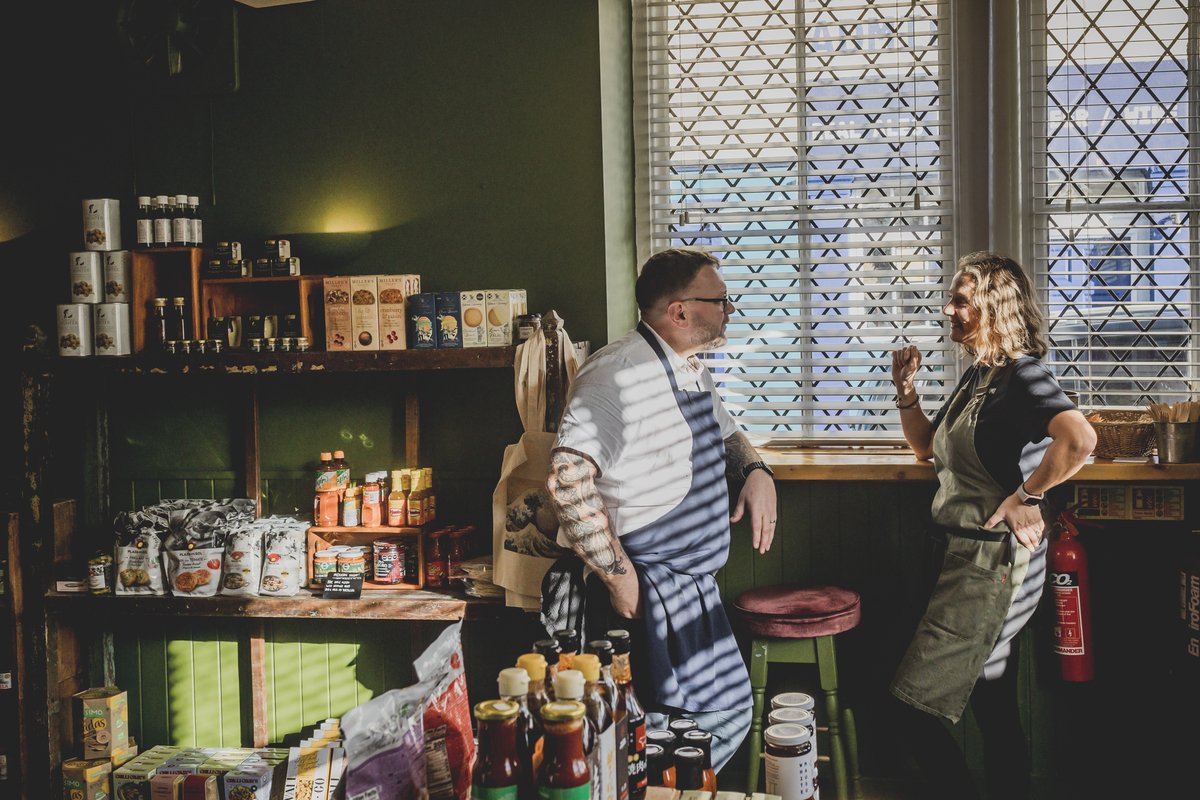 Two people in aprons talking in a store with shelves of packaged food products and a window with blinds.