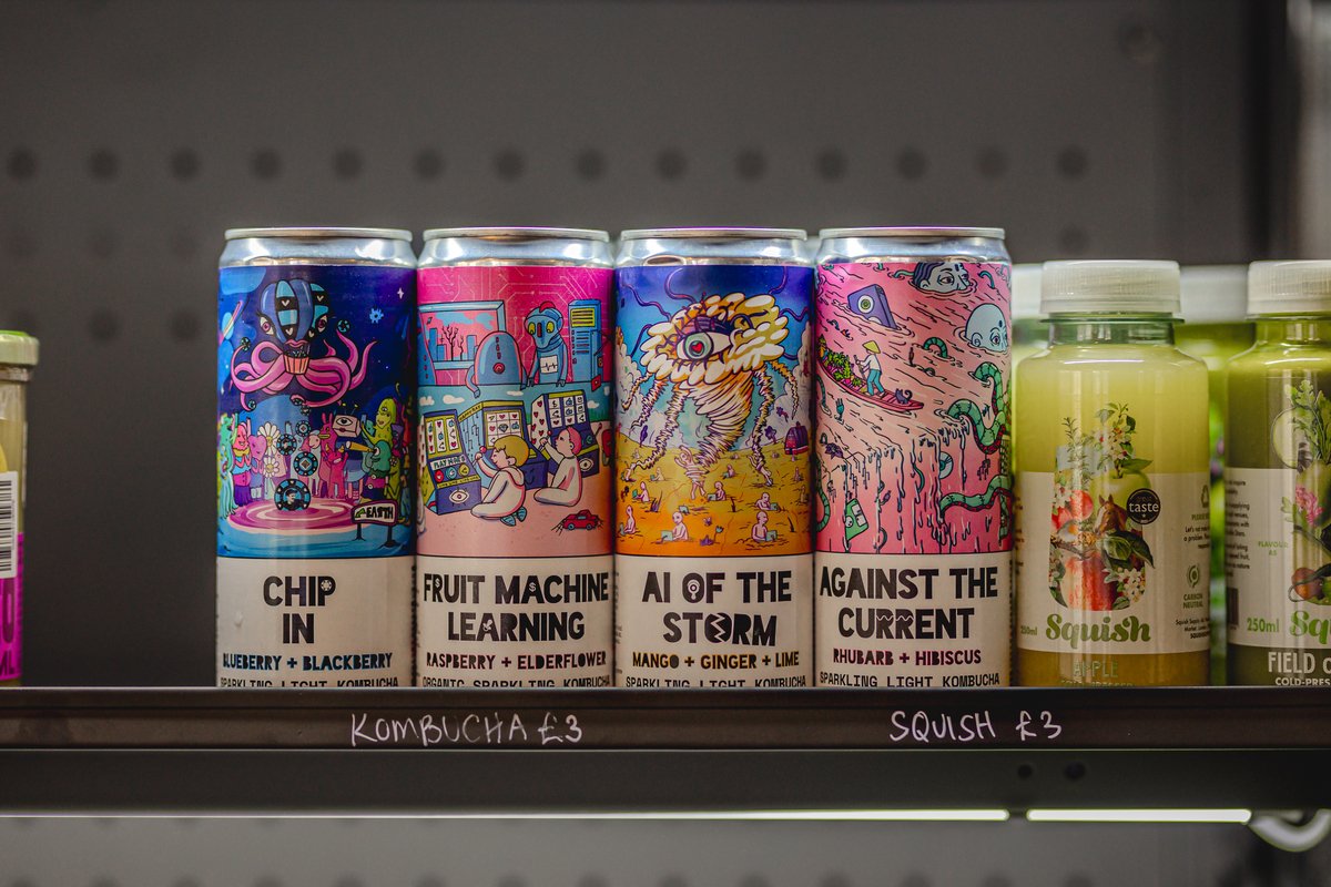 Assorted cans of kombucha with colorful, artistic labels and flavors like blueberry, raspberry, mango, and rhubarb on a shelf, alongside bottled drinks labeled 'Squish.'