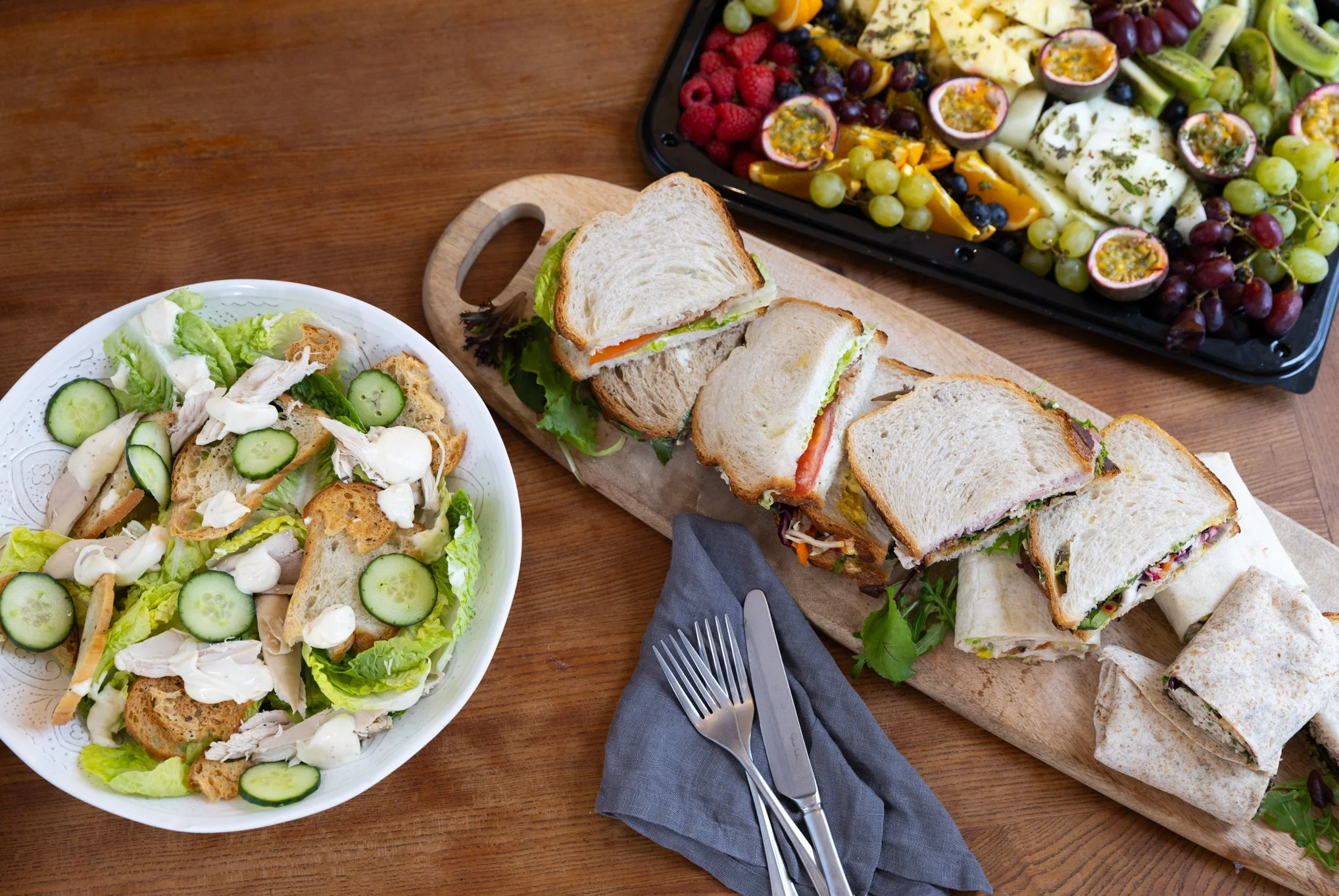 A spread of food on a wooden table including a fresh green salad with cucumbers, croutons, and shredded chicken in a white bowl; a platter of assorted sandwiches with various fillings on a wooden board; and a tray of assorted fresh fruits like grapes