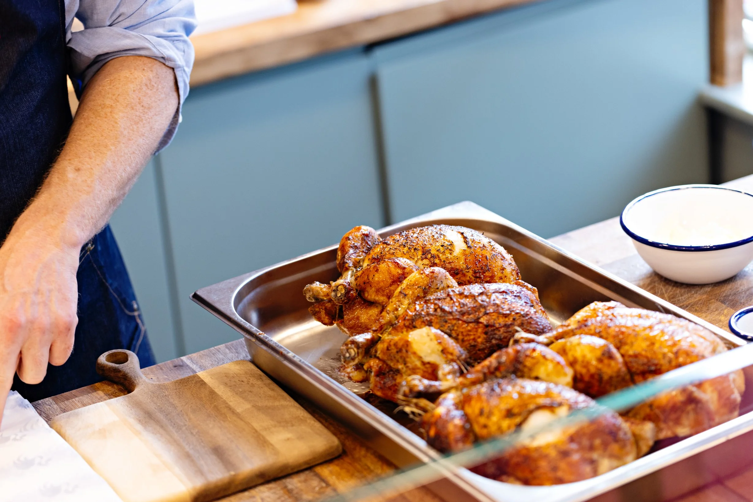 Several cooked chickens in a metal tray on a wooden table, with a person nearby and a white bowl with a blue rim in the background.