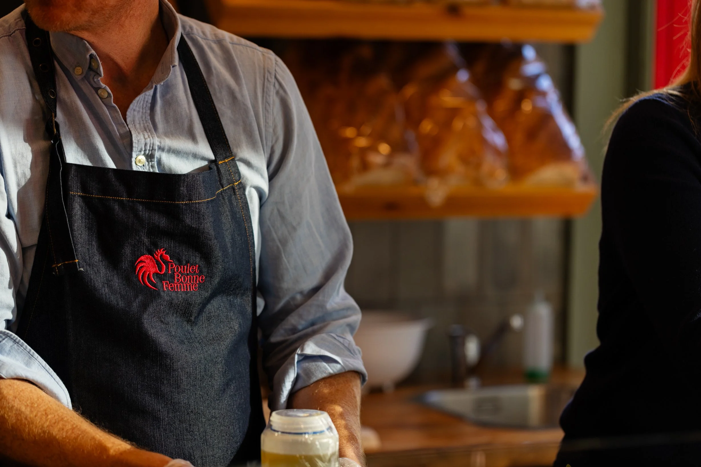 Person wearing a dark apron with a red rooster logo and French text, standing in a kitchen with jars and a sink in the background.