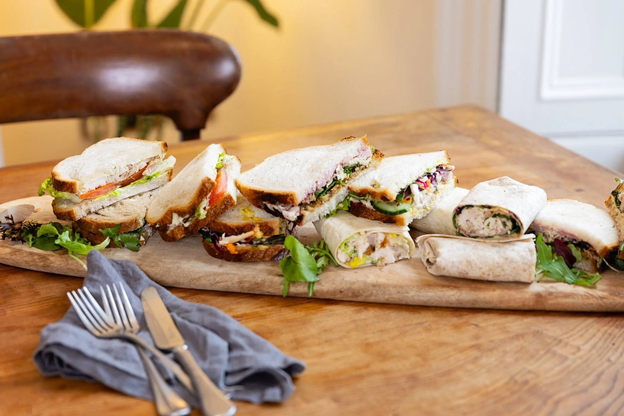 Assorted sandwich halves on a wooden table with a fork and knife on a gray napkin in the foreground.