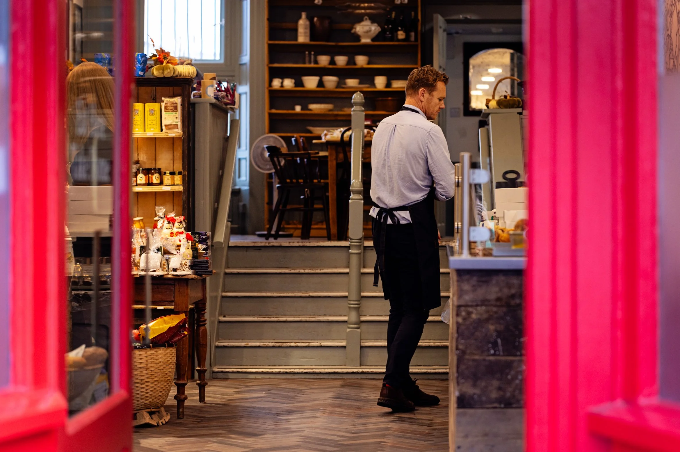 A man in a white shirt and black apron working behind the counter of a shop or cafe, seen through pink window or door frames.
