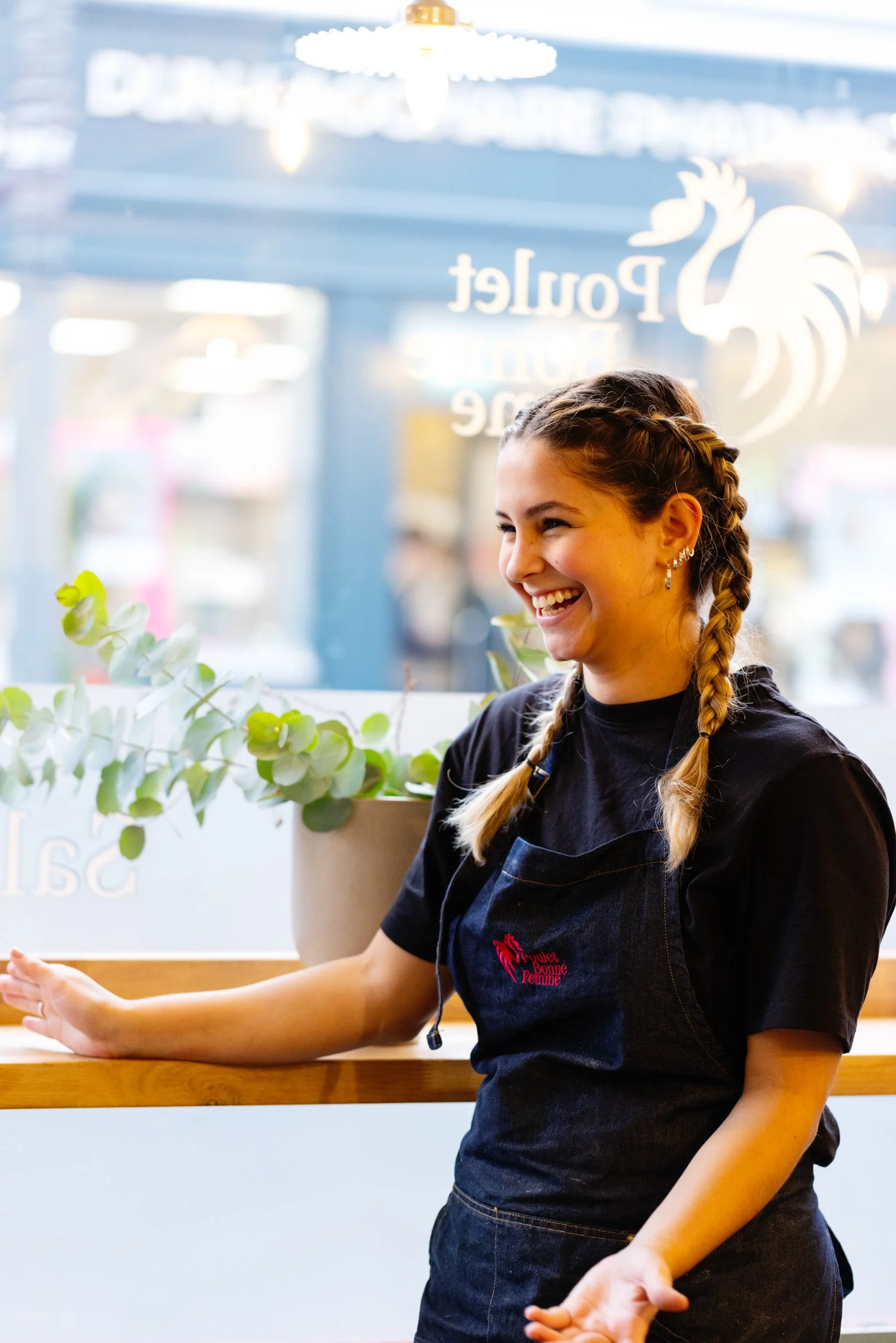 A smiling young woman in a black apron with dainty earrings inside a cafe or restaurant, with a window behind her displaying the name 'Poulet' and a rooster logo, and a plant on the counter.