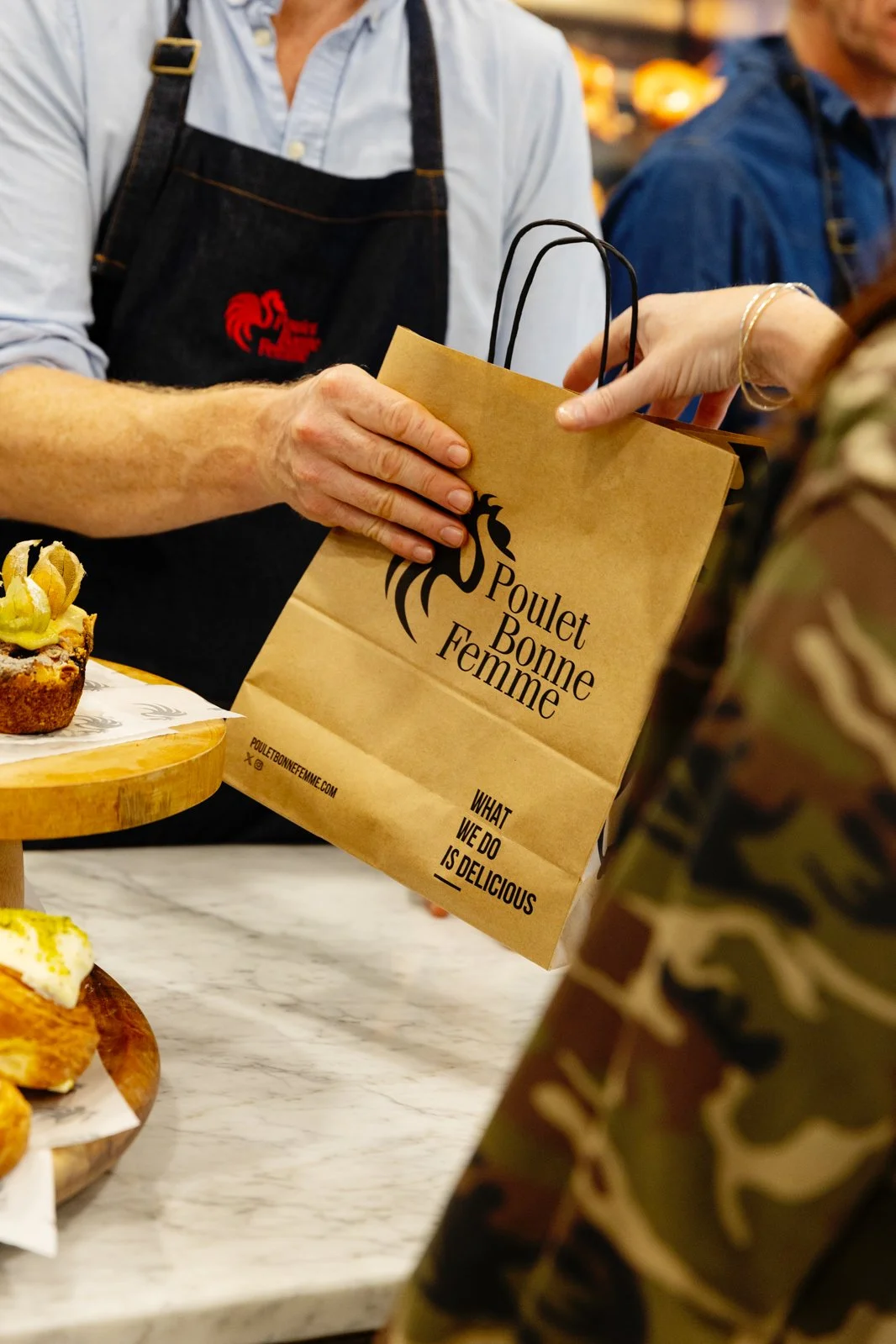 A person in an apron handing a paper bag to another person at a food stand. The bag has the logo and name "Poulet Bonne Femme" on it, along with the slogan "What we do is delicious." There are pastries on the table, and another person in a camouflage