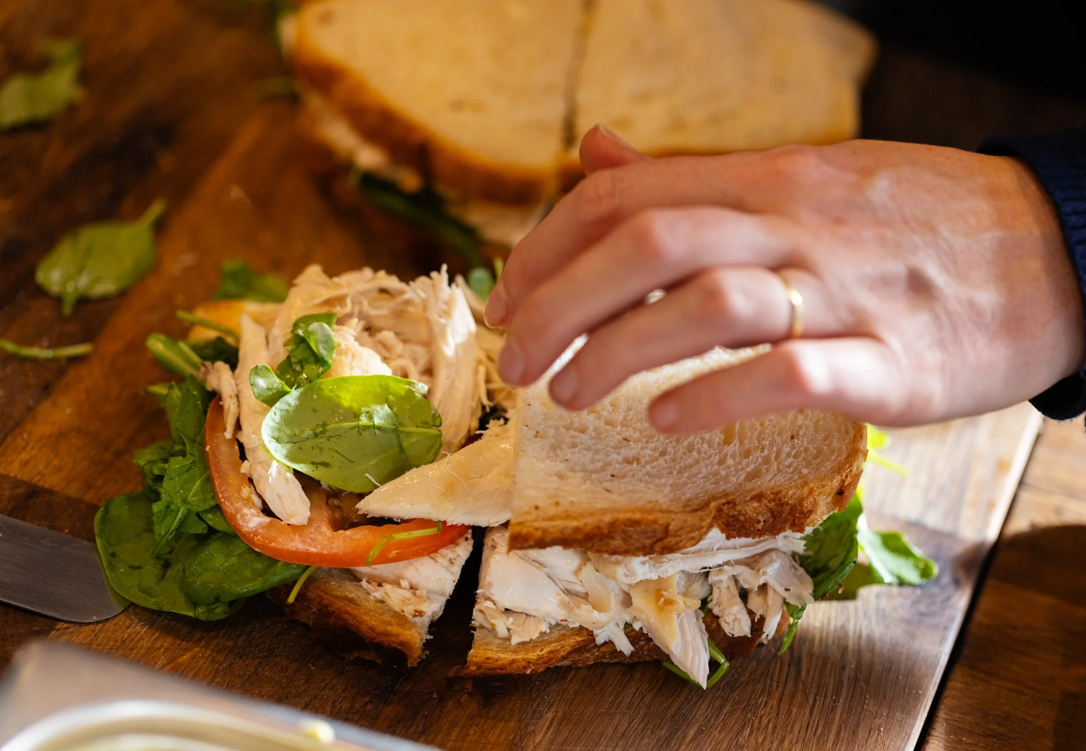 A person preparing a sandwich with shredded chicken, lettuce, tomato, and cheese on whole wheat bread on a wooden cutting board.