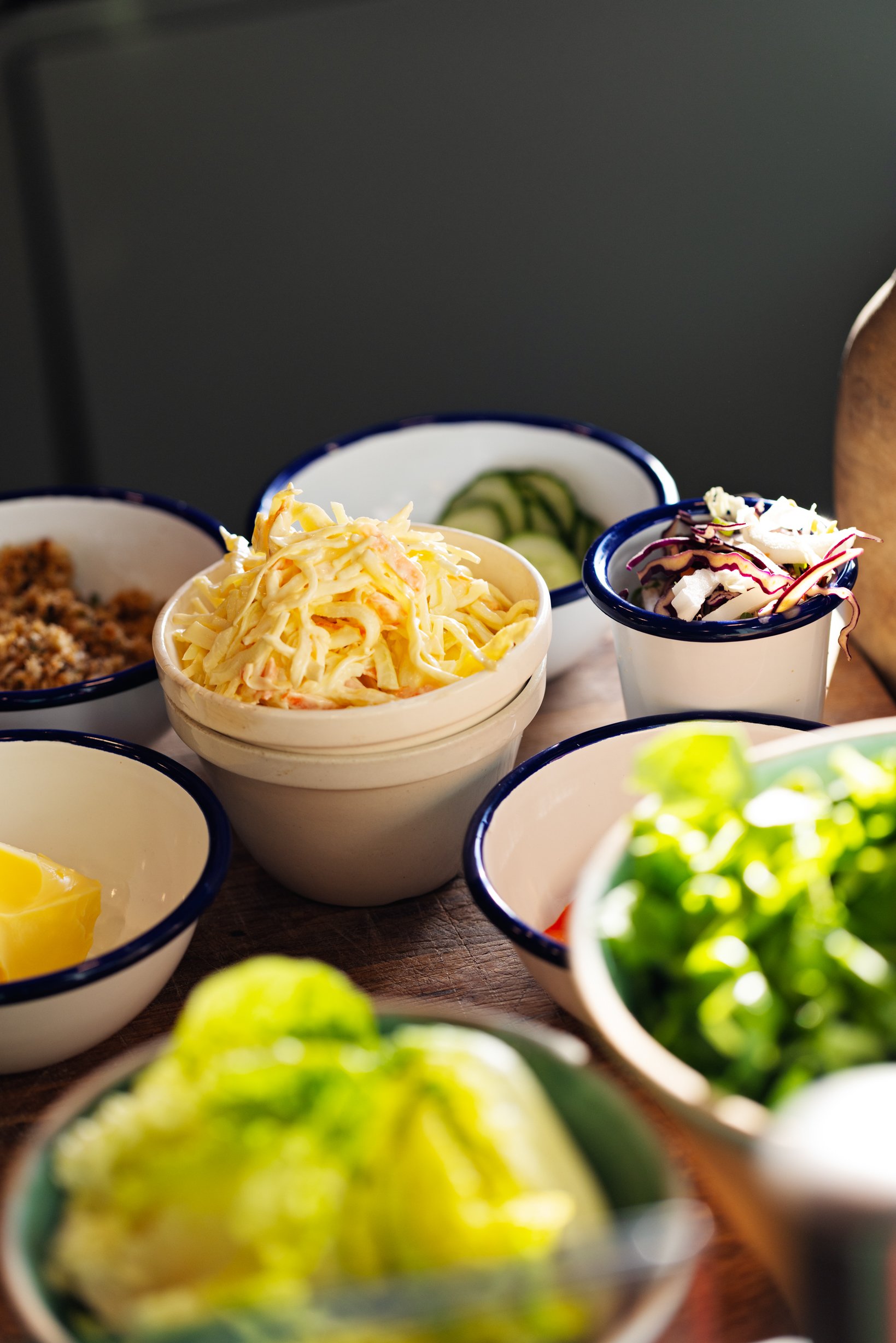 Assorted bowls of salad, coleslaw, cucumber slices, and other ingredients on a wooden table.