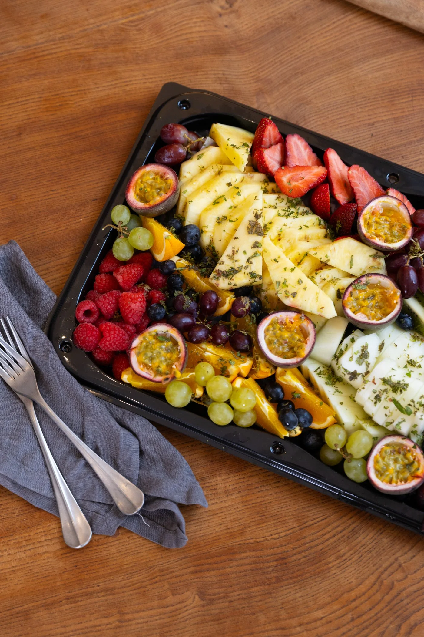 Assorted fresh sliced fruits including strawberries, pineapples, grapes, passion fruit, blueberries, raspberries, and oranges on a black serving tray with forks on a gray napkin.
