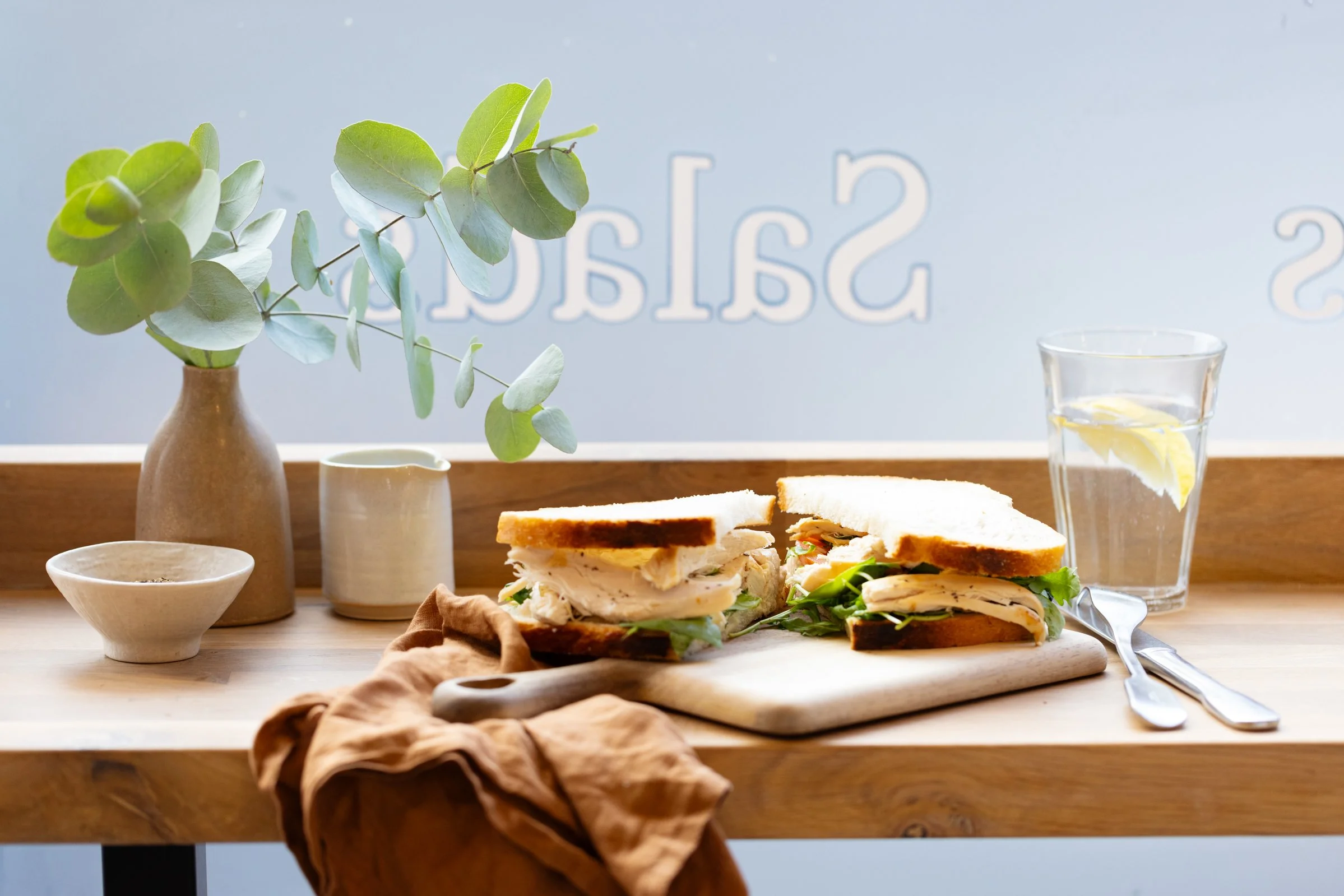 A sandwich on a wooden cutting board with lettuce and turkey, a glass of lemon water, a potted eucalyptus plant, and some bowls and utensils on a wooden table against a light blue background.