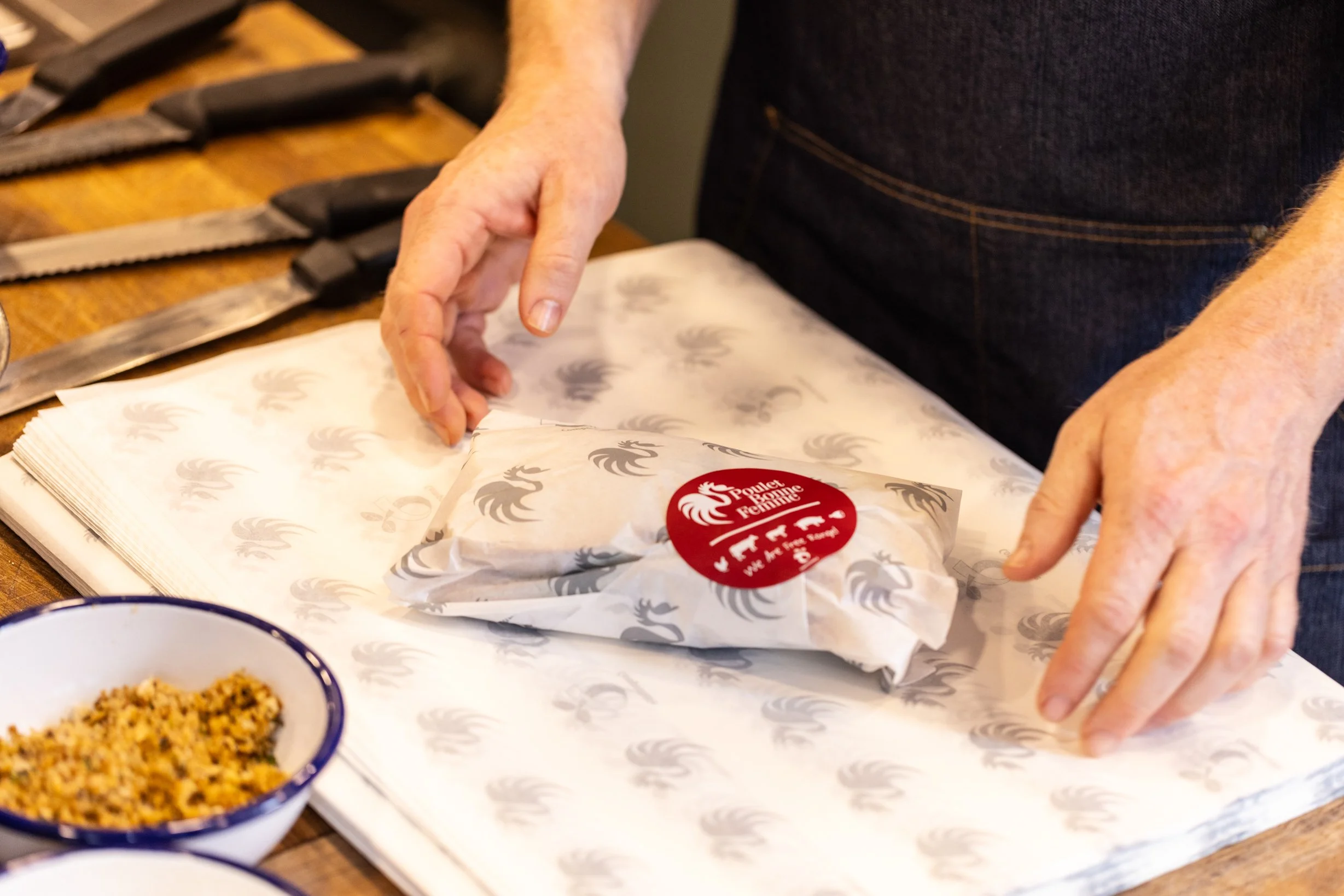 Person wrapping a package with branded paper that has a rooster logo, on a table with a bowl of seasoned rice and knives nearby.