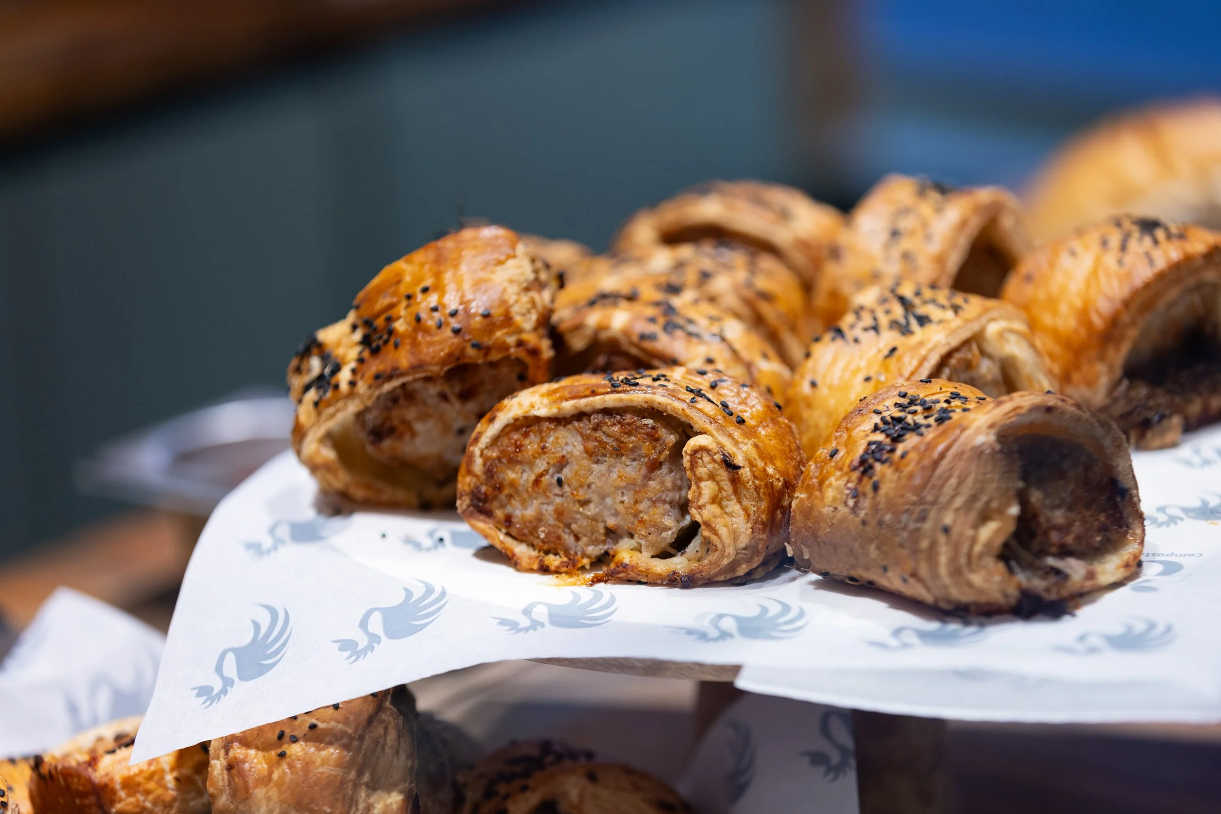 Close-up of sausage rolls with golden-brown, flaky pastry, topped with black sesame seeds, arranged on white paper with blue logos.