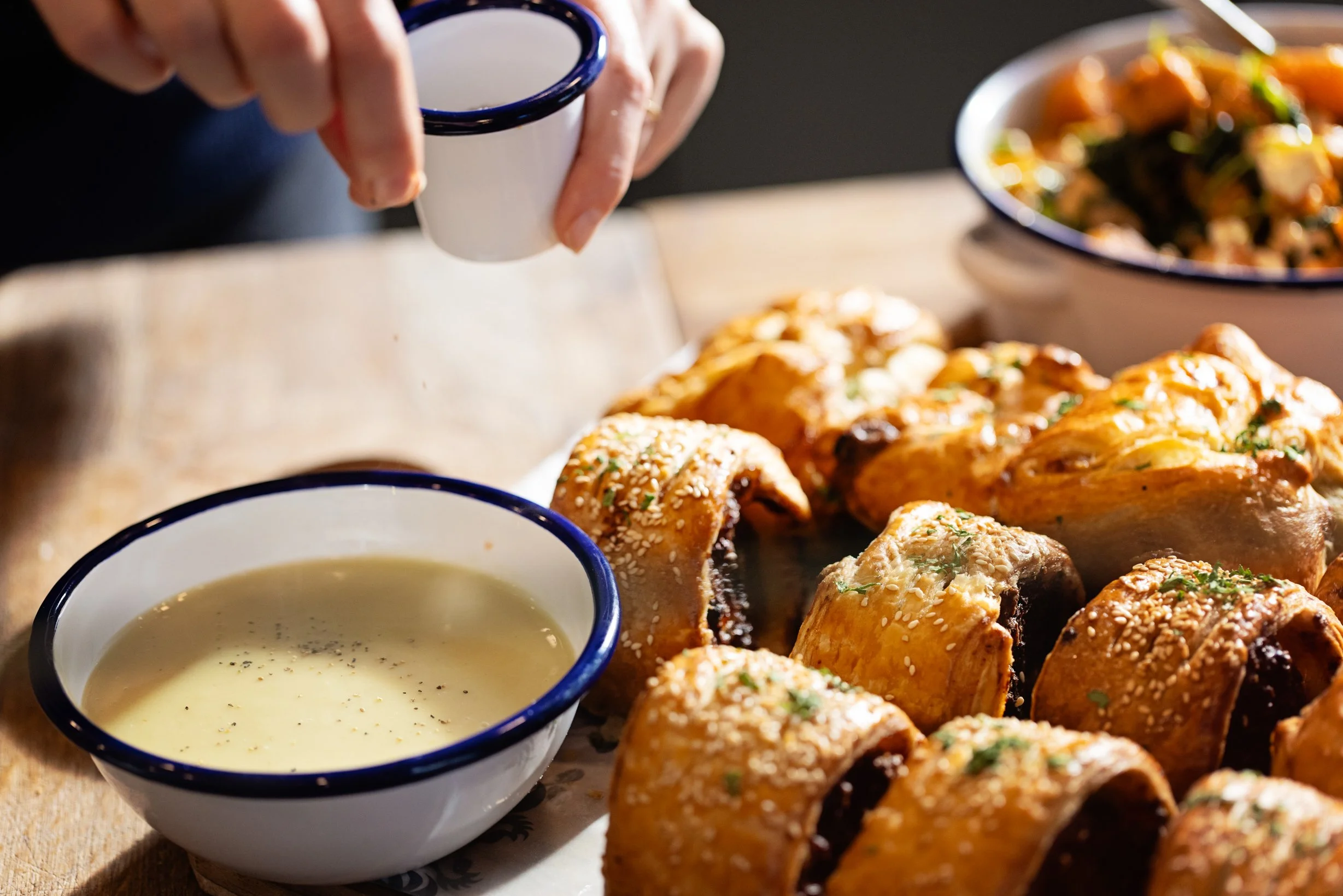 A hand sprinkling salt on baked bread rolls with sesame seeds on top, along with a bowl of creamy soup in the foreground and a bowl of mixed vegetables in the background.