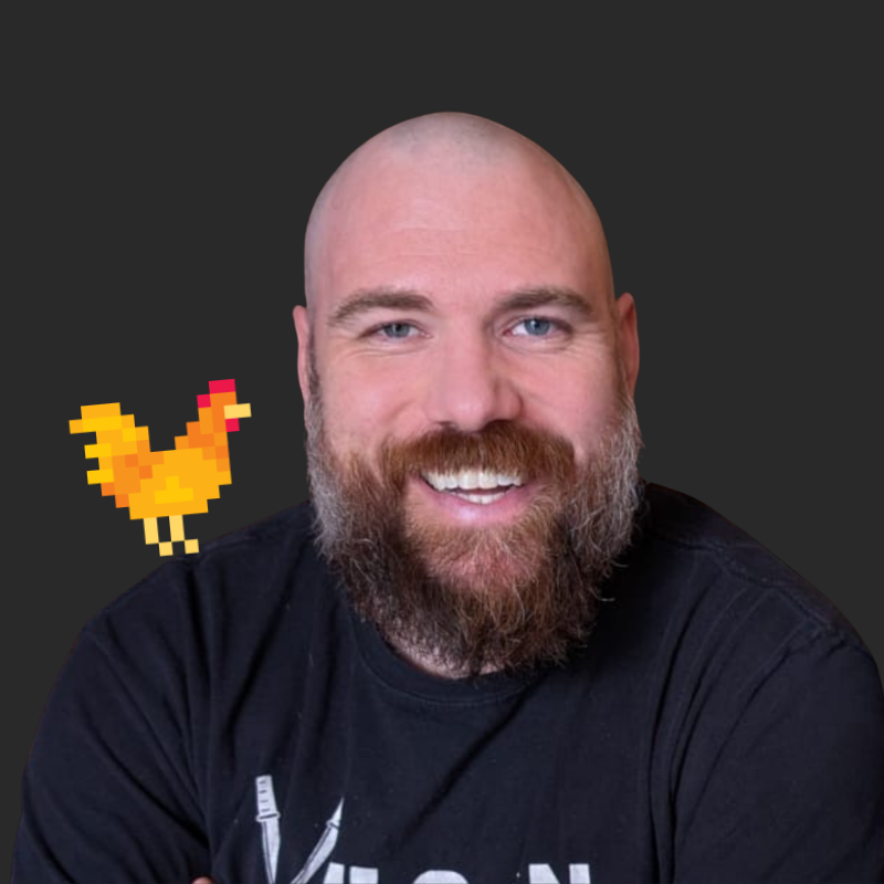 A smiling man with a beard and bald head, wearing a black T-shirt, against a dark background with a pixelated chicken graphic on his shoulder.