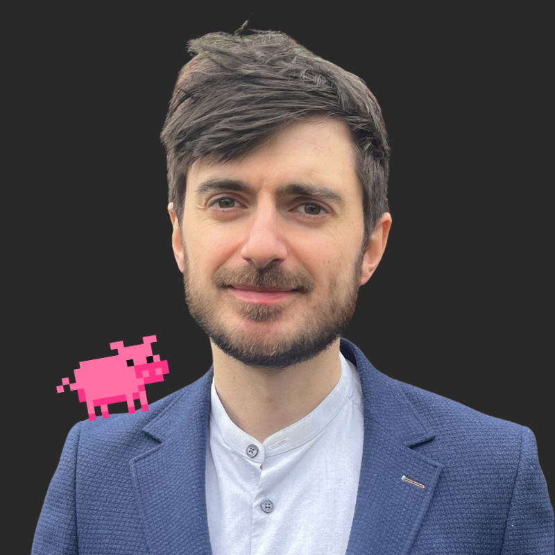 A young man with brown hair, blue eyes, a beard, wearing a blue blazer and white shirt, smiling at the camera with a pixel art pink pig on his shoulder against a black background.