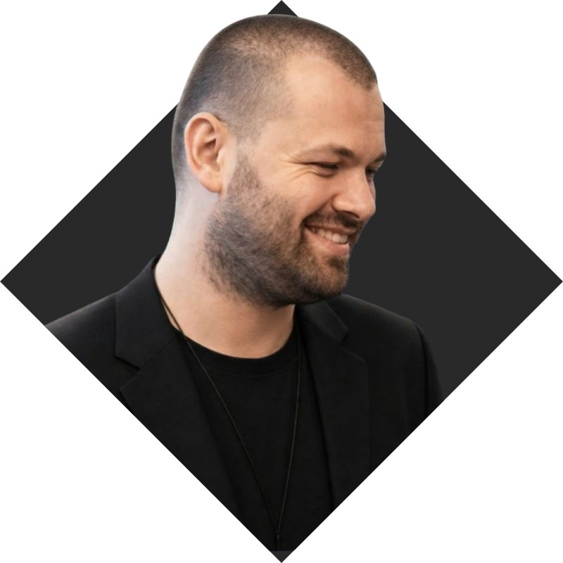 A smiling man with a short beard and buzz cut, wearing a black blazer and black shirt, on a dark geometric background.