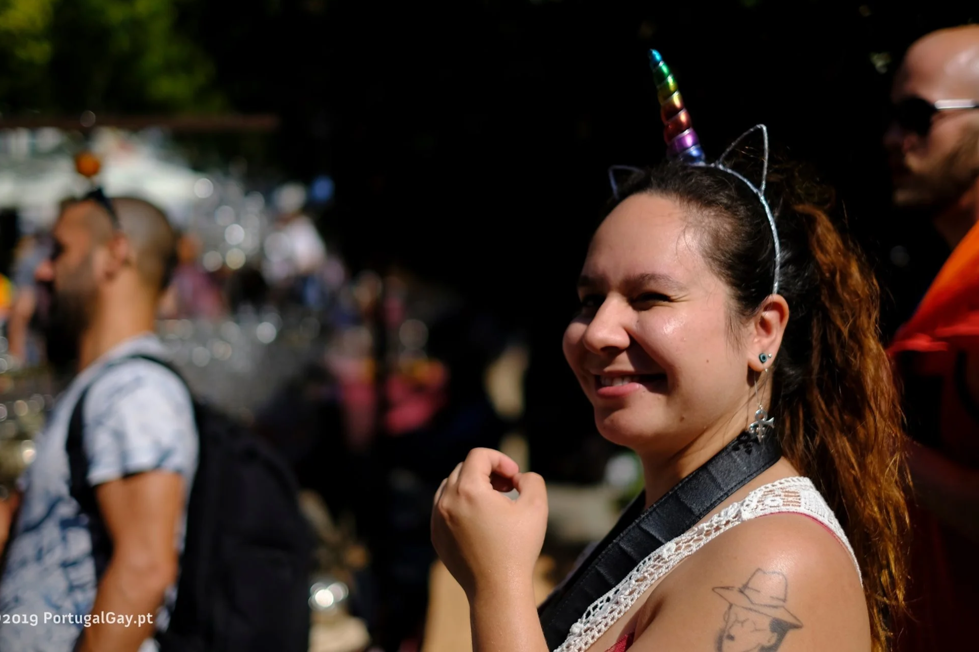 A woman with a rainbow unicorn headband and cat ear headband, smiling and winking at the camera at Pride event in Lisbon, with people and booths visible in the blurry background.