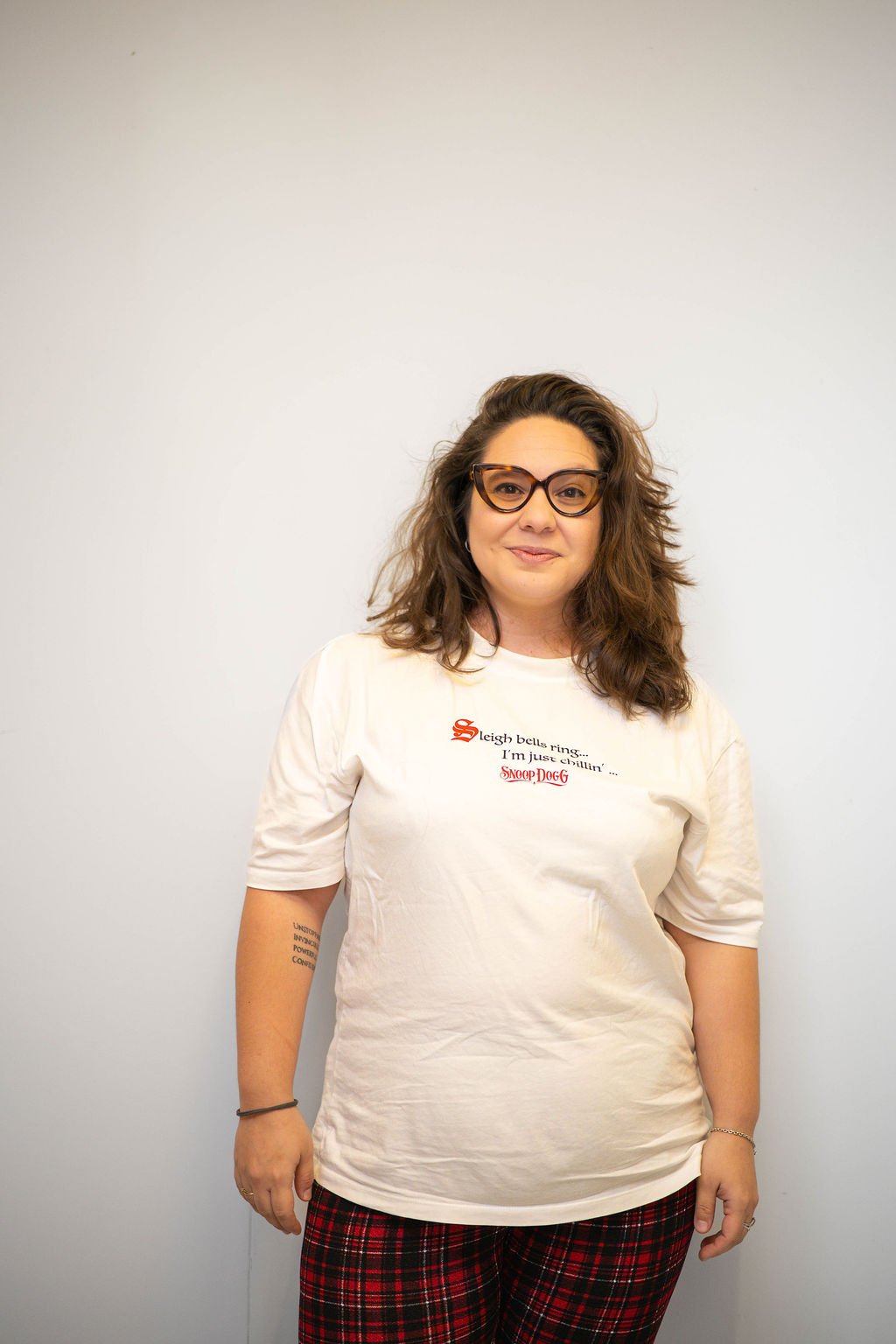 Woman with glasses and long curly hair wearing a white t-shirt with printed text and red plaid pants, standing against a plain white wall.