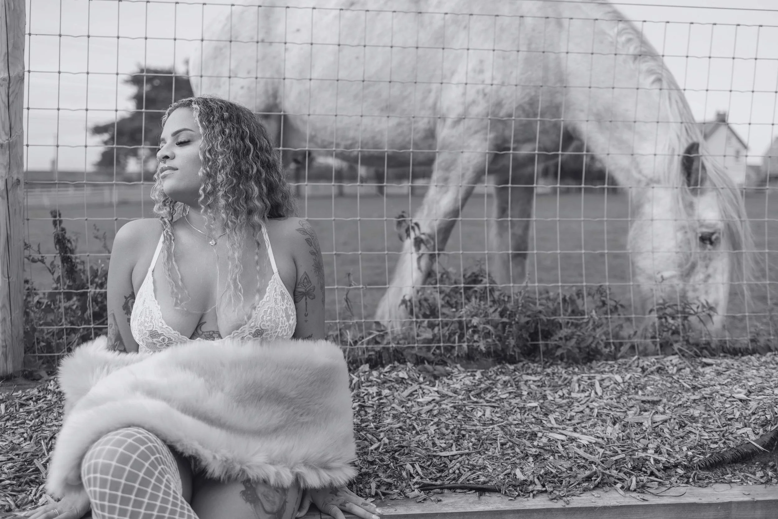 A woman with curly hair and tattoos sitting outside near a fenced area with a white horse, in a black-and-white photograph.