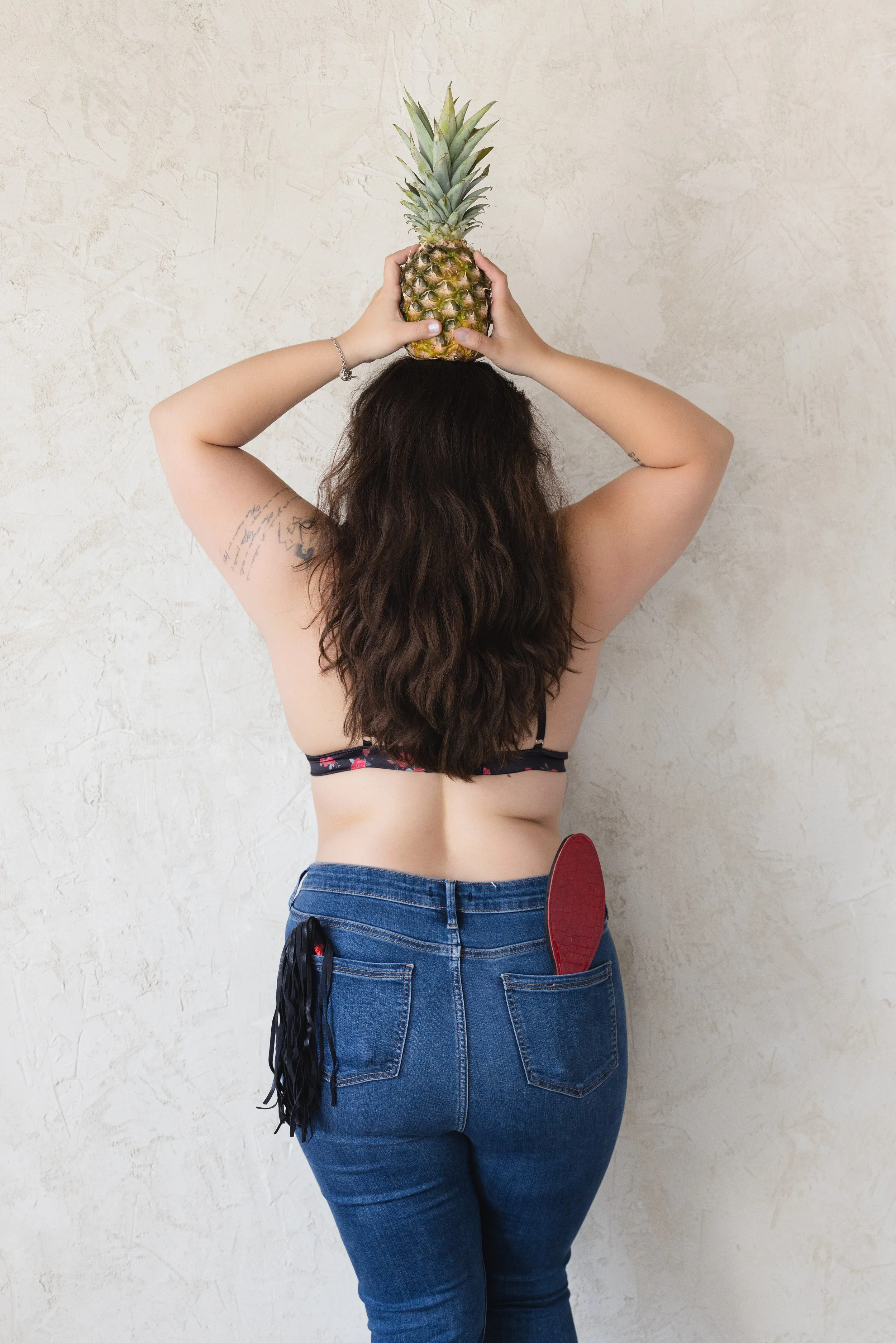 A person with long dark hair wearing a black spaghetti strap top and blue jeans, standing against a plain textured wall. They are holding a pineapple on their head with both hands, and a red shoe is visible in their back pocket and a black tassel hanging from the pocket.