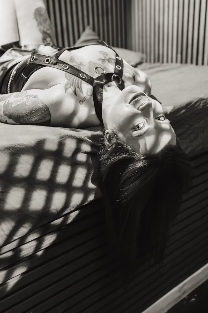 A woman with tattoos lying upside down on a bed, with shadows cast on her body from a patterned object above.