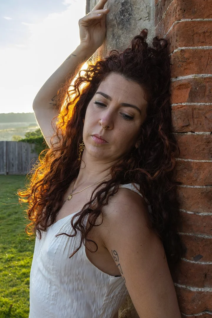 Young woman with curly red hair and tattoos, leaning against a brick wall outdoors at sunset, looking contemplative.