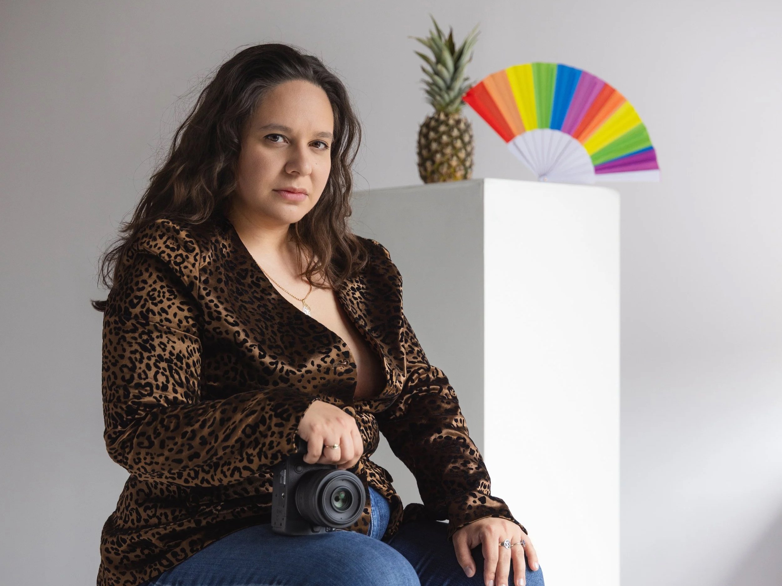 A woman with long curly hair wearing a leopard print top holding a camera, sitting against a plain gray wall with a pineapple and colorful paper fan in the background.