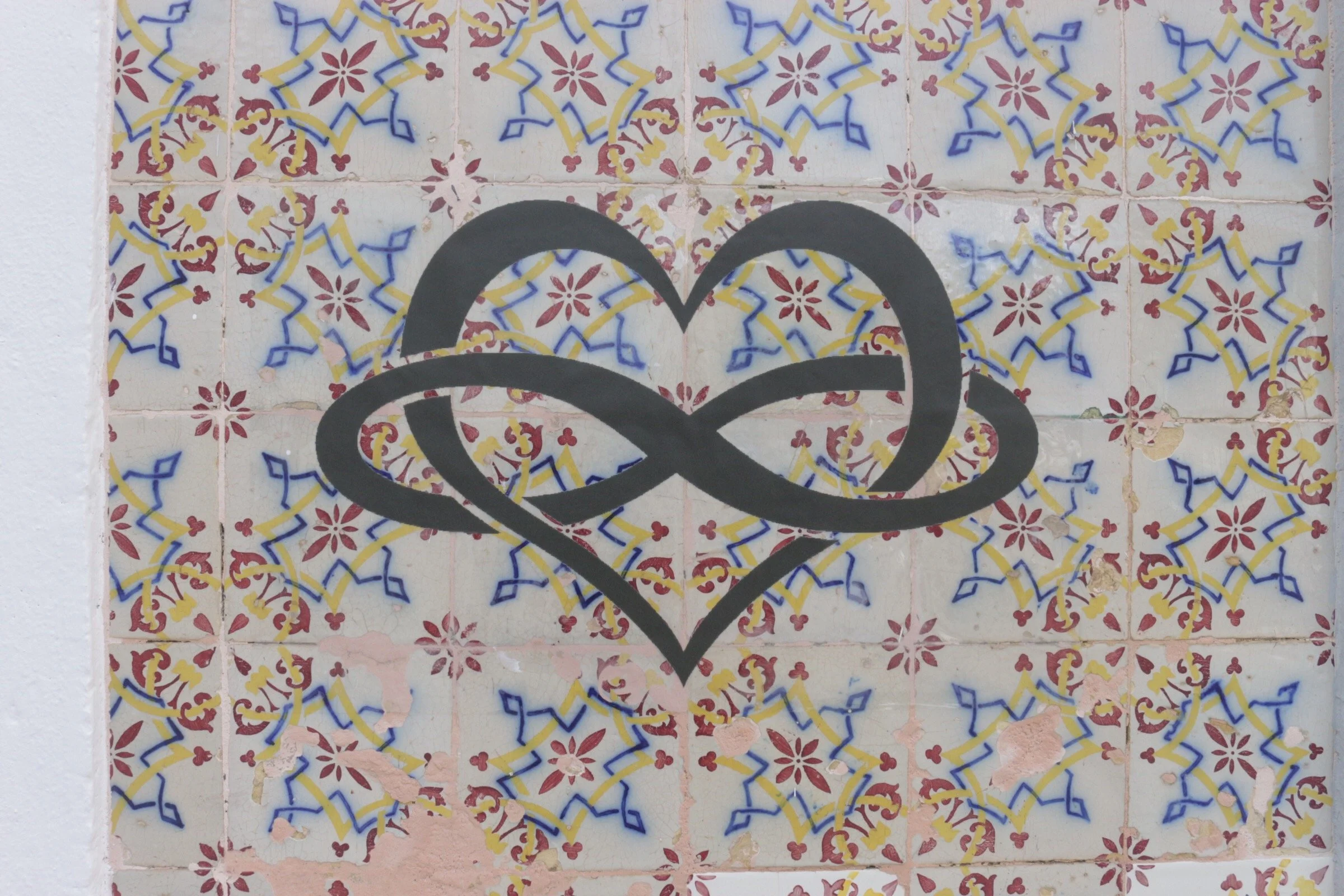 A black outline of a heart intertwining with a swirl is painted over a colorful patterned tile background. Logo of Ana Elisa Baptista's Documentary Film & Social Research Project ‘The Poly-Womxn Perspective on Polyamorous Relationships in Portugal'