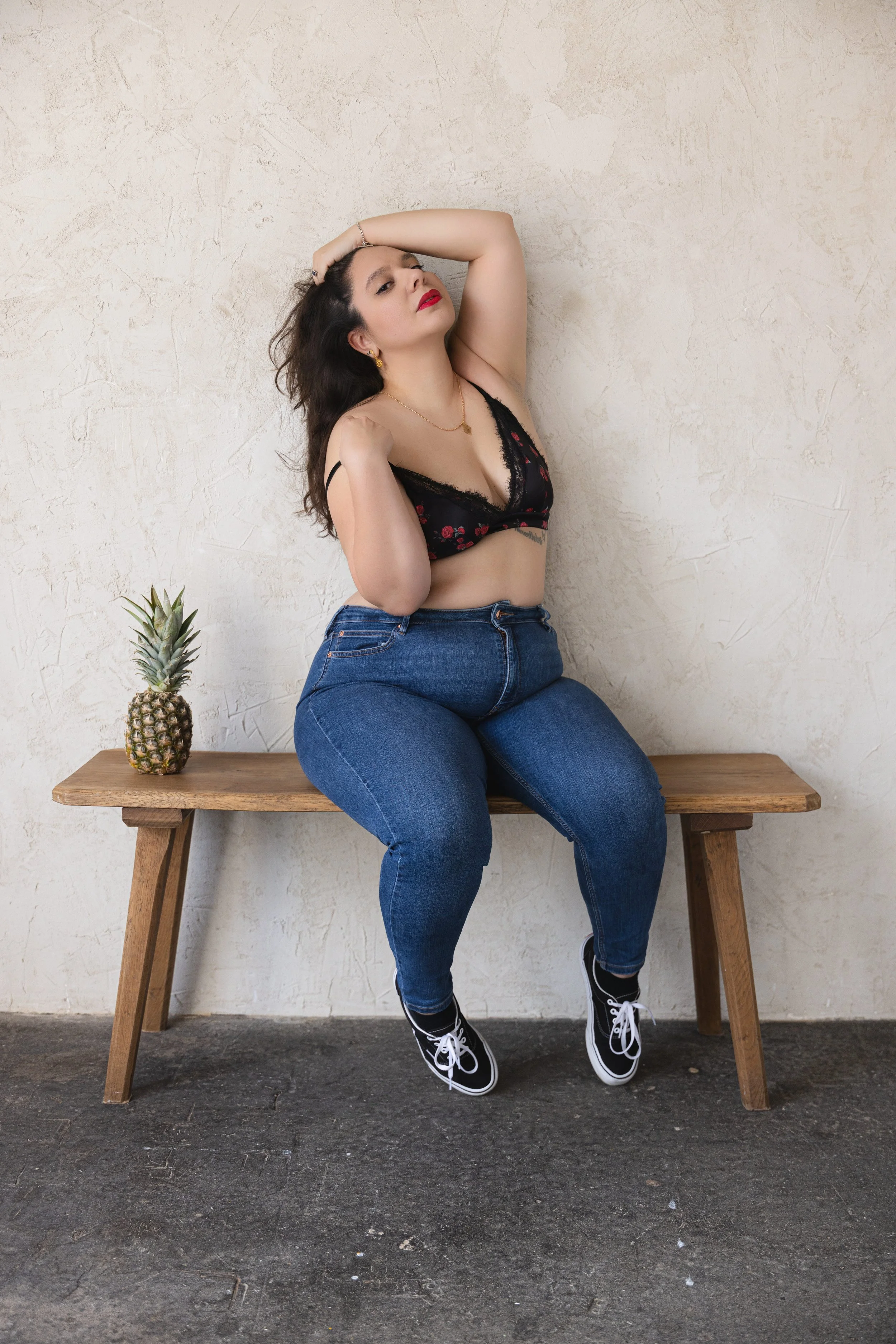 A woman with dark hair and red lipstick is sitting on a wooden bench against a beige wall. She is wearing a black lace floral crop top, blue jeans, and black sneakers. One hand is resting on her head while the other is touching her shoulder. There is a pineapple placed on the bench beside her.